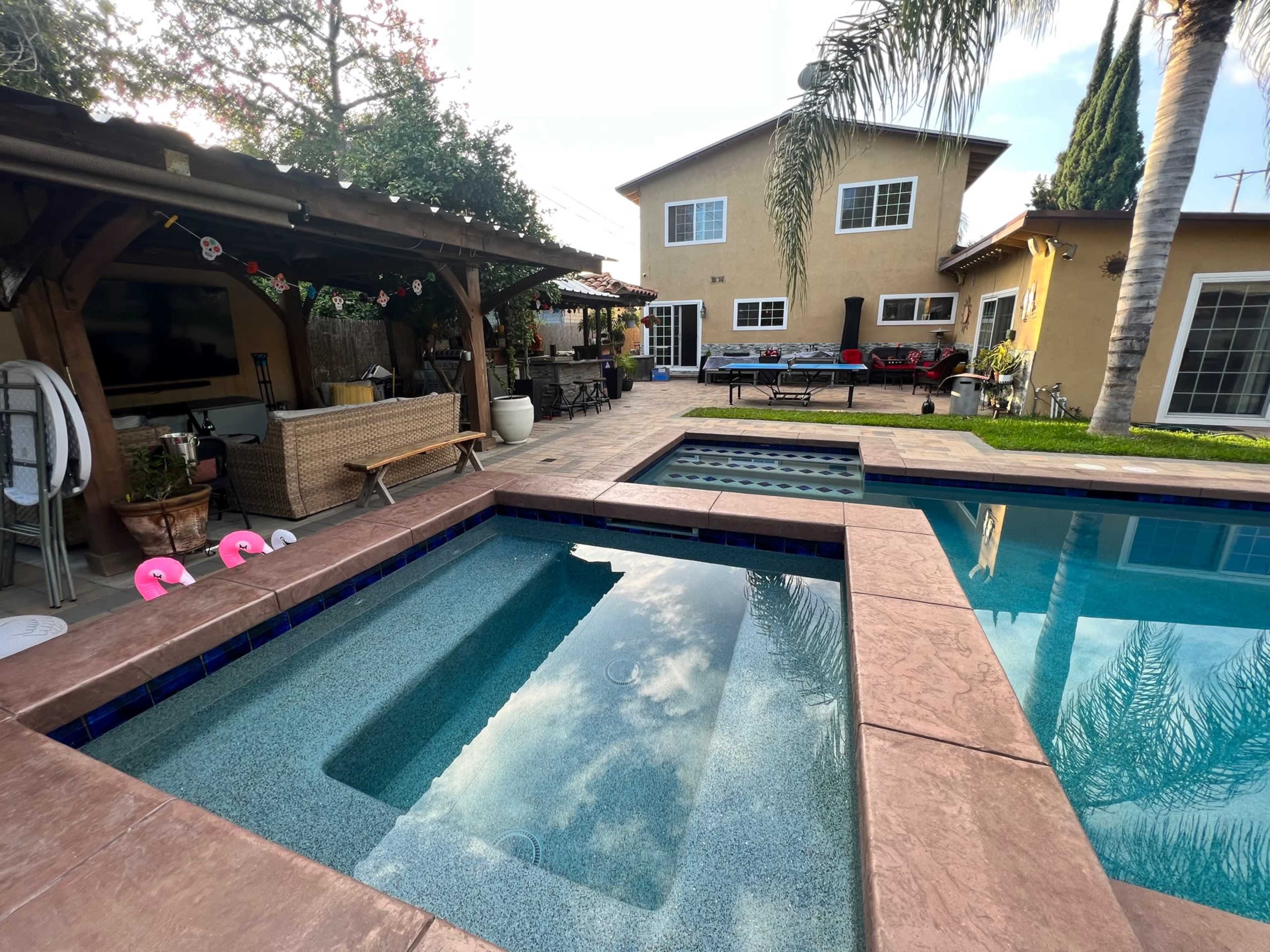 A backyard features a swimming pool with a spa, surrounded by concrete decking and a two-story house in the background.