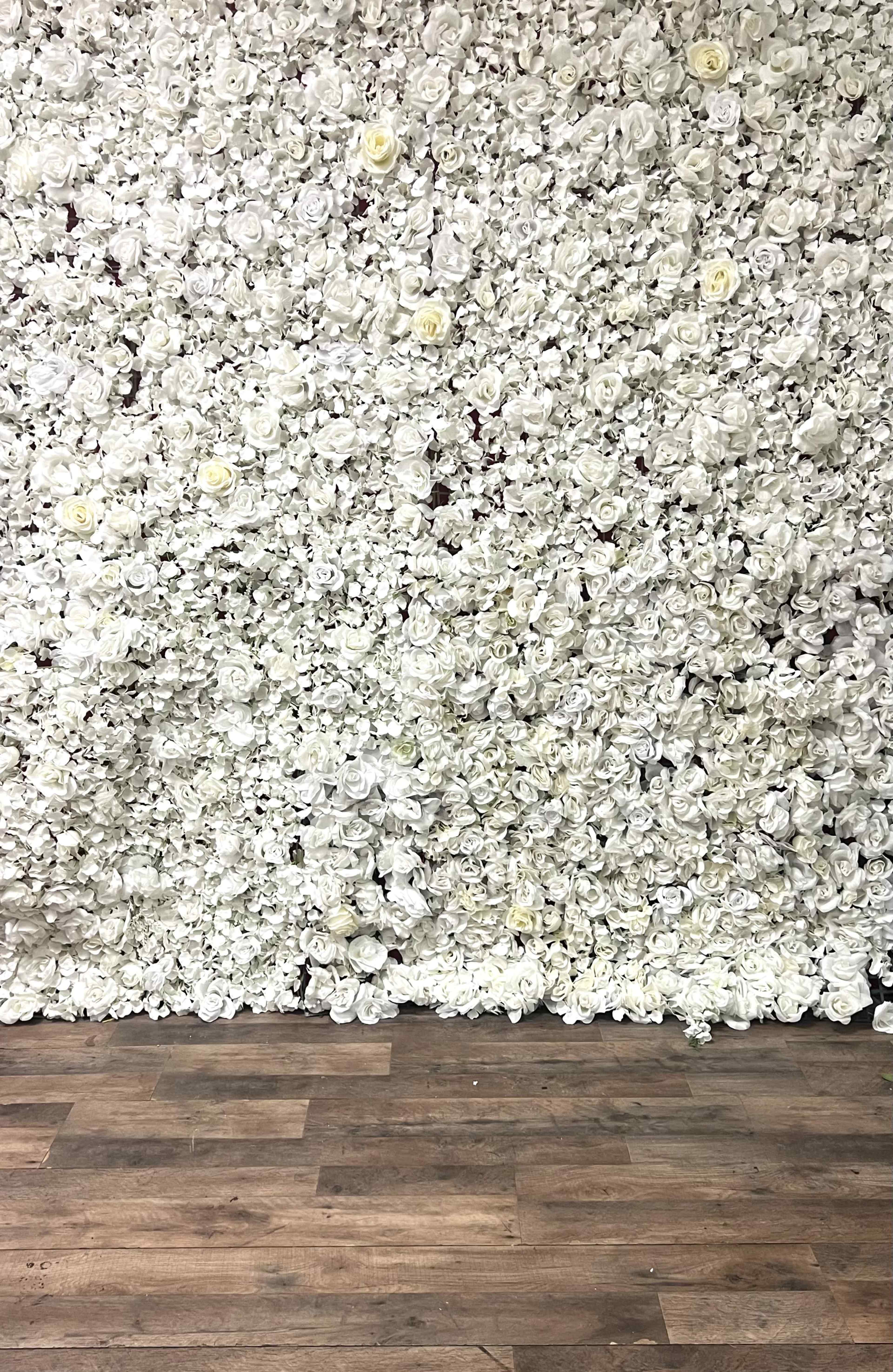 The image shows a wall covered entirely in artificial white roses, with a wooden floor visible at the bottom.