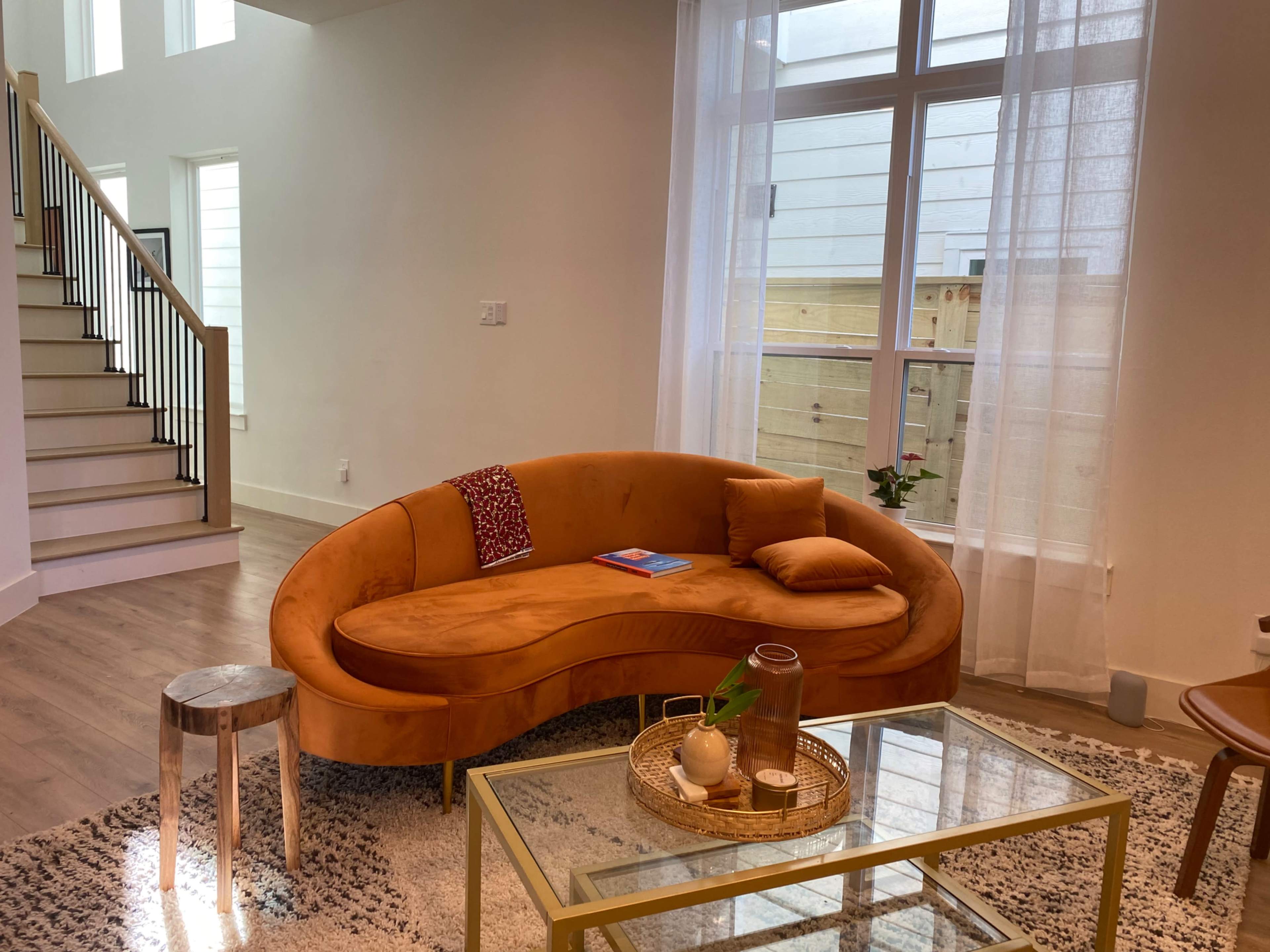 A curved orange sofa is positioned next to a glass coffee table and a small wooden stool in a bright, modern living room with large windows.