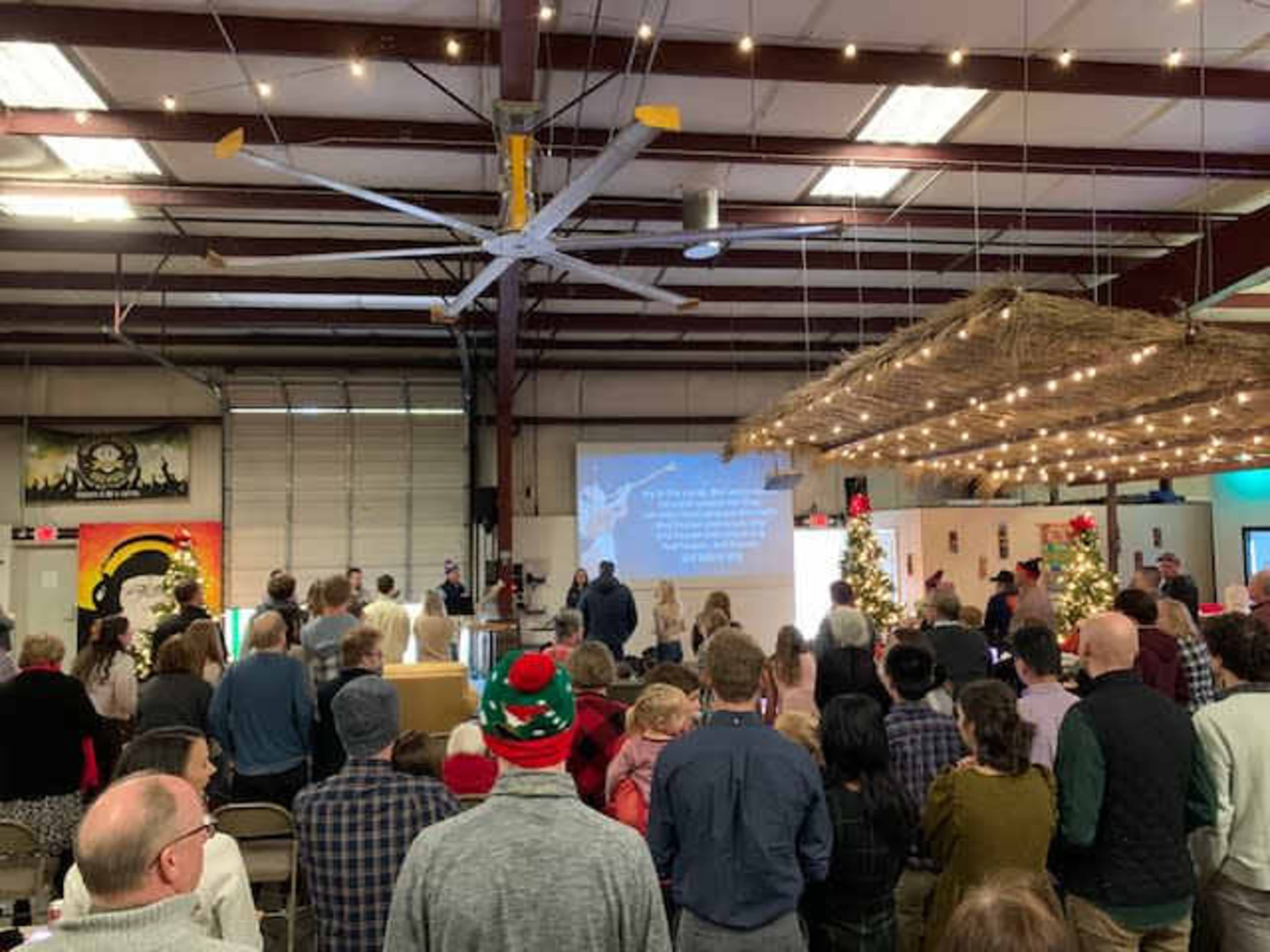A large group of people is gathered in a warehouse for a festive event, with a speaker and a projected presentation seen in the background.