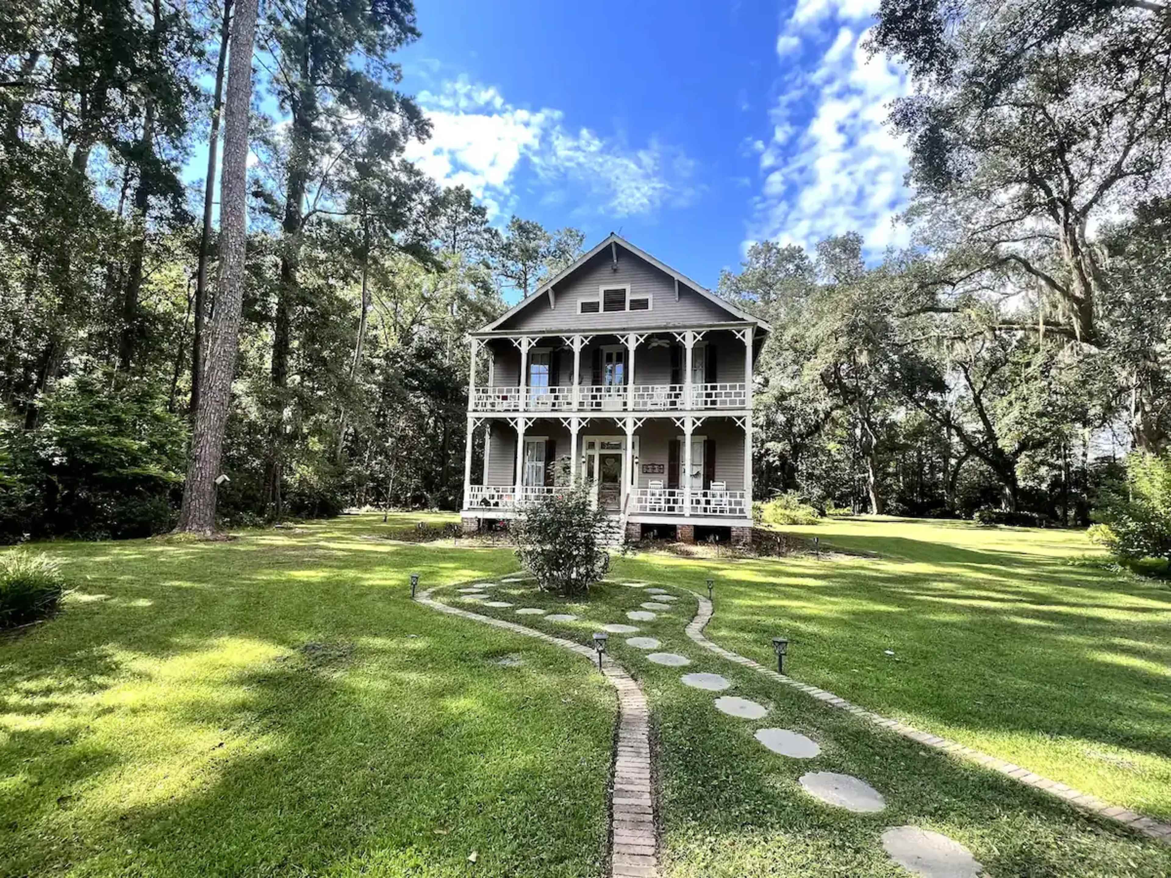 A two-story house with a wraparound porch is set in a landscaped yard surrounded by trees.