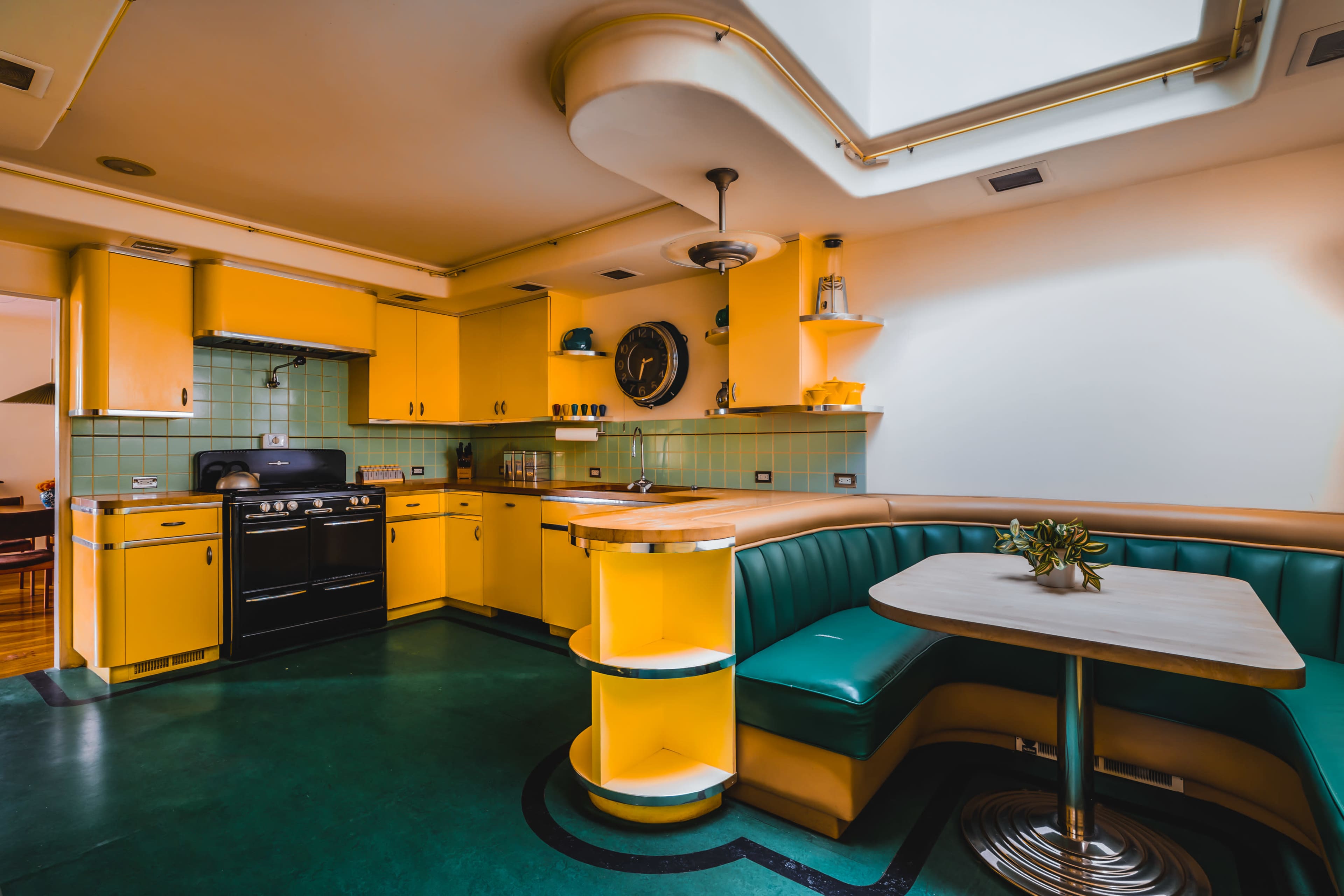 The image shows a retro-style kitchen with yellow cabinetry, a green booth, and a round table set in a corner.