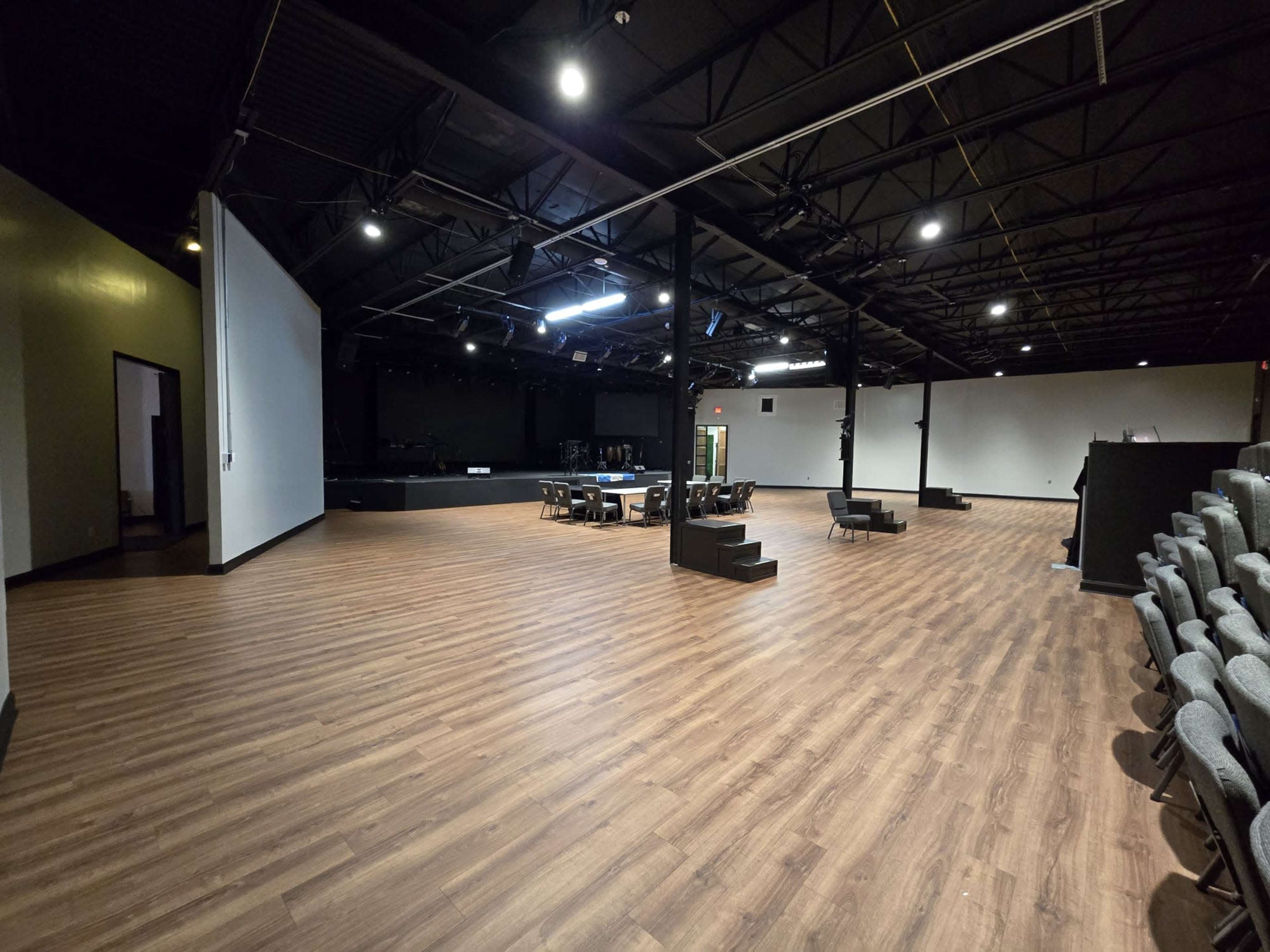 The image shows a spacious, well-lit performance venue with a wooden floor and seating areas, featuring a stage at the far end.