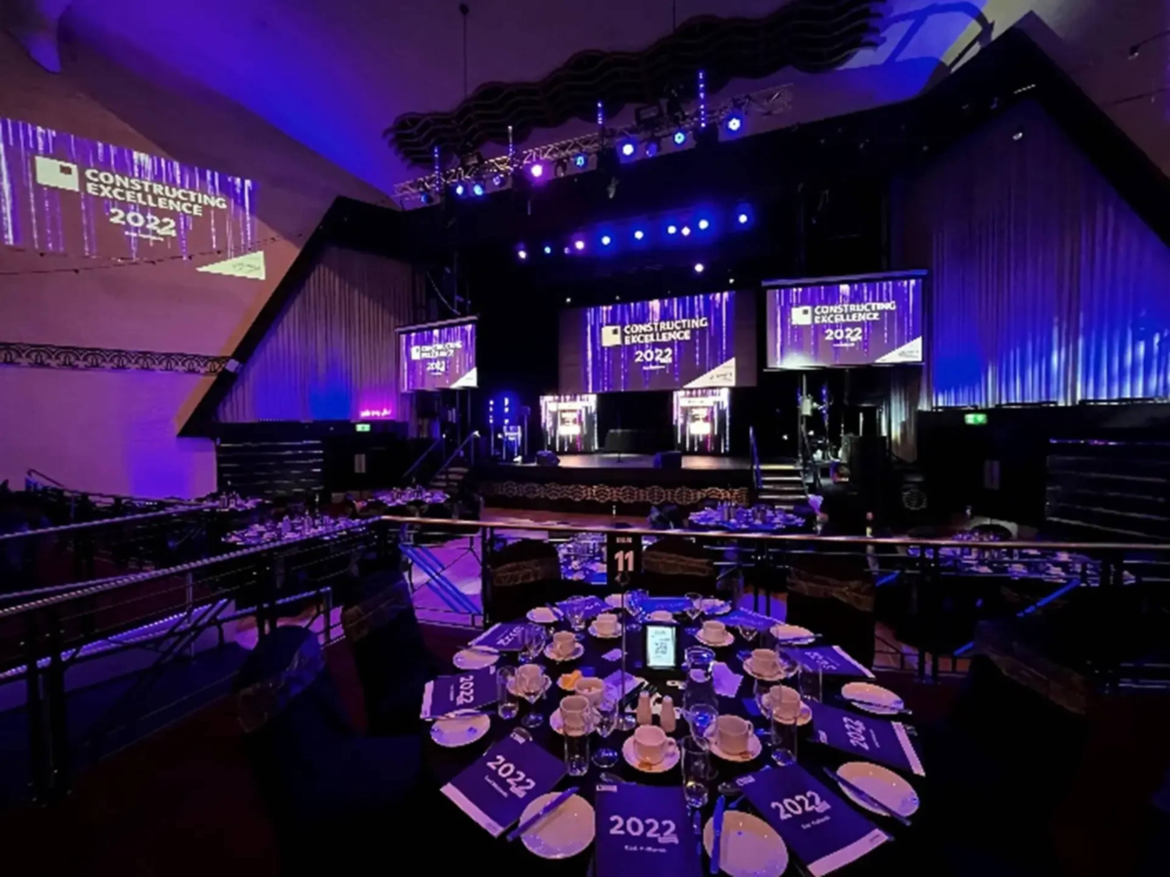 The image shows a banquet setup with tables elegantly arranged, illuminated by colorful stage lights and featuring large screens displaying "Construction Excellence 2022."