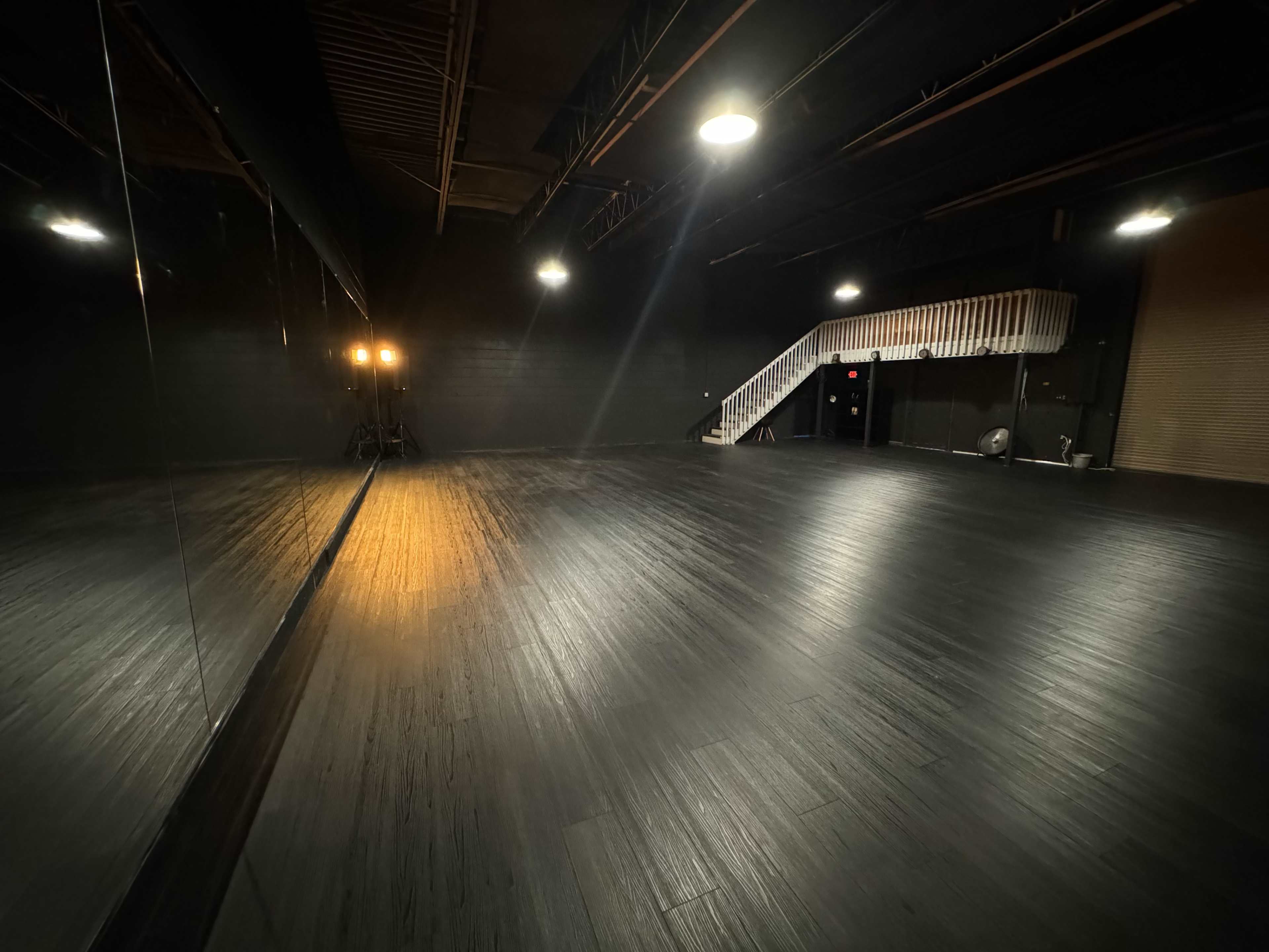 The image shows a dimly lit dance studio with a polished black floor, illuminated by overhead lights and featuring a staircase in the background.