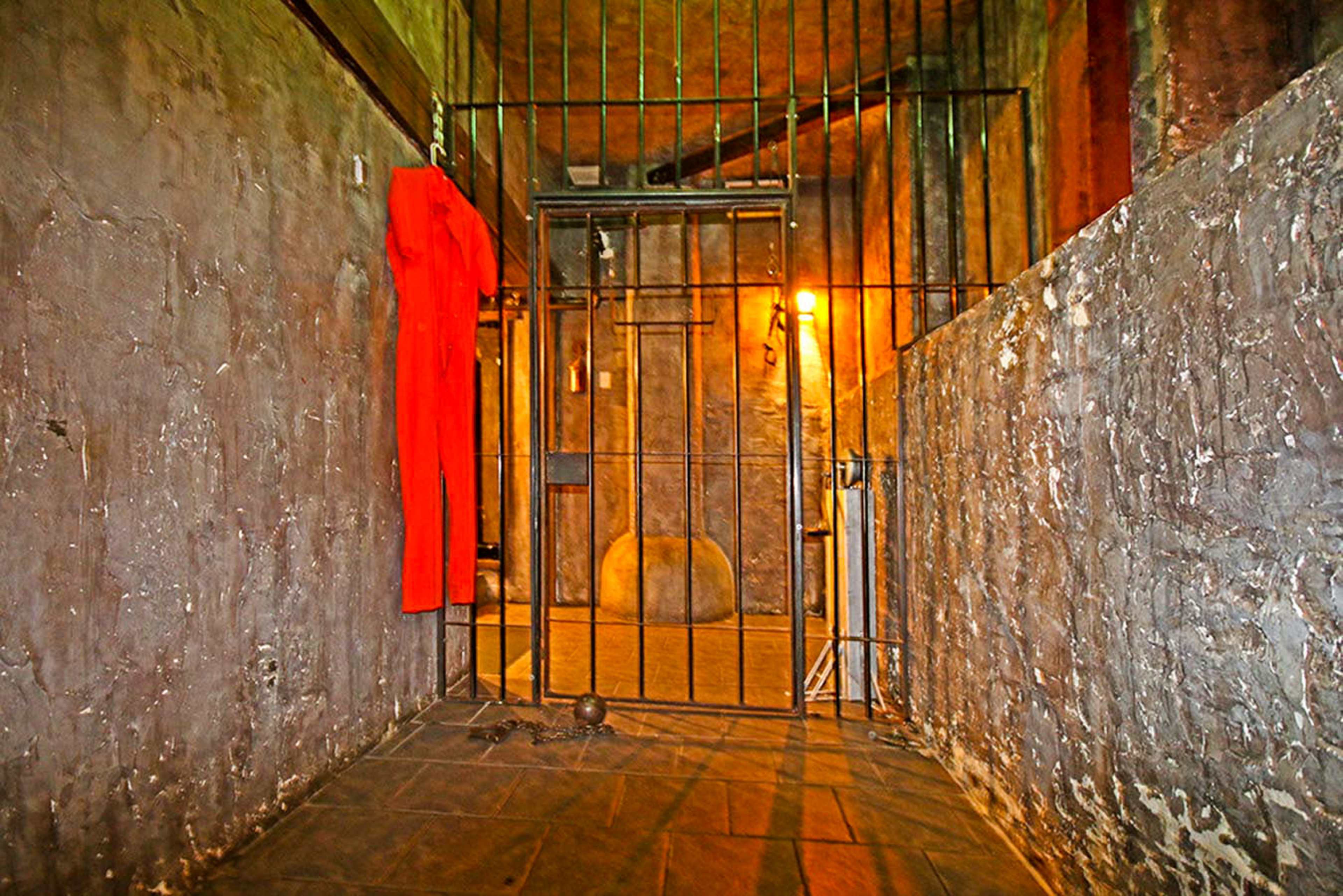 The image shows a dimly lit prison cell with a barred gate and a red prison jumpsuit hanging nearby.