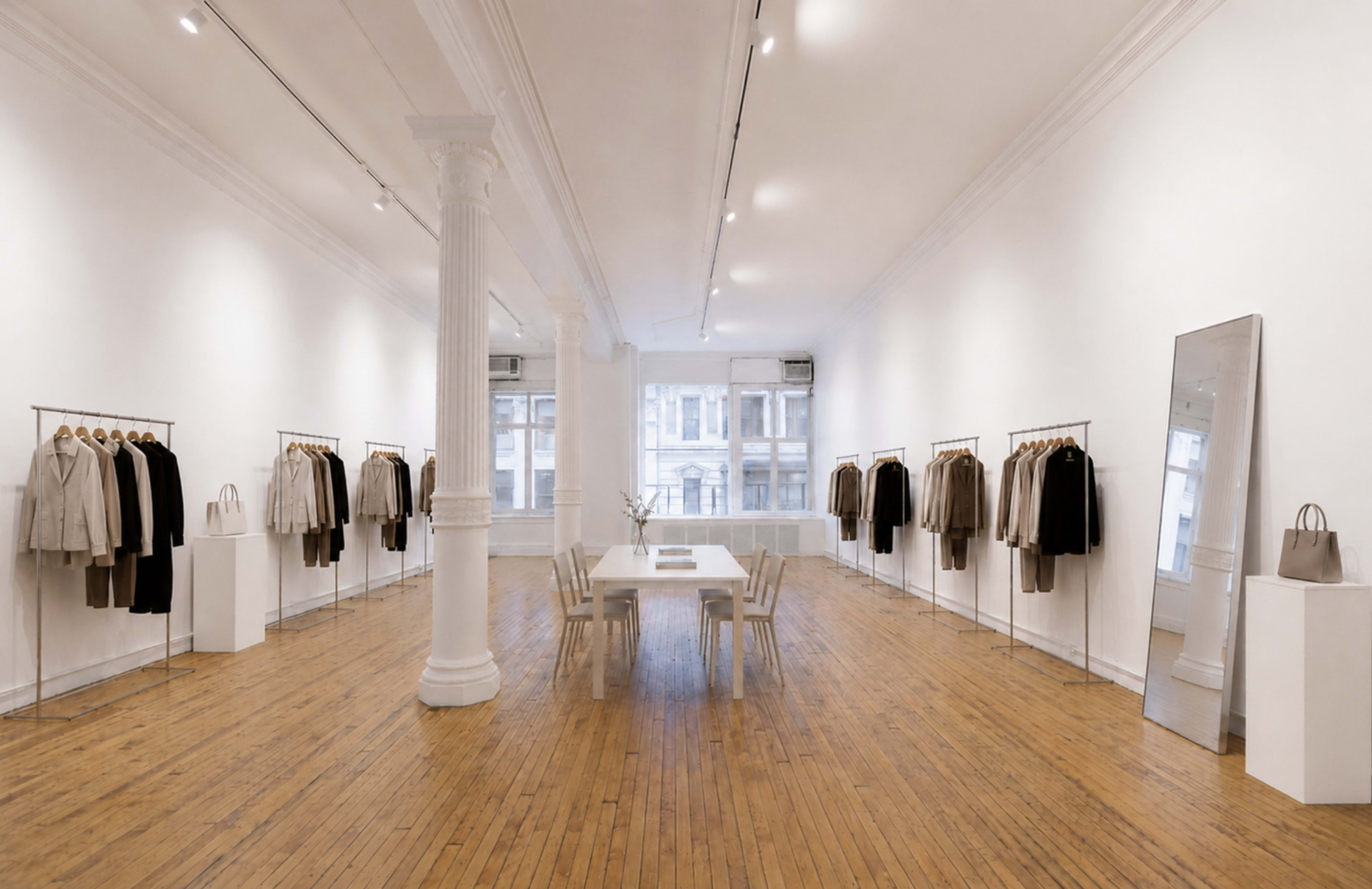 The image shows a spacious art gallery with white walls, wooden flooring, and clothing displayed on racks along the sides, featuring a central table surrounded by chairs.