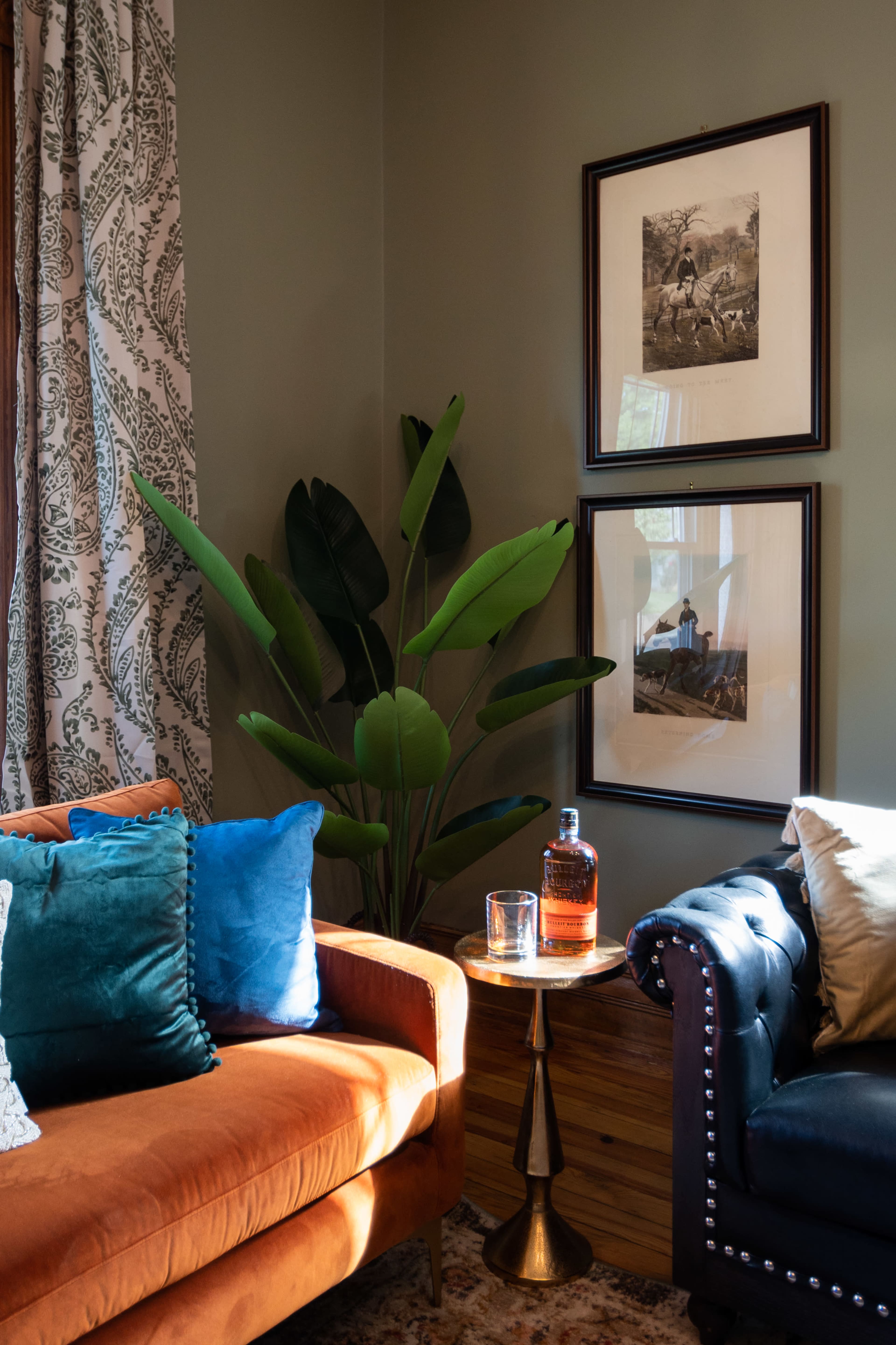 A cozy living room corner features a brown couch, a blue couch, a small side table with a bottle and glass, and framed artwork on the wall beside a large plant.