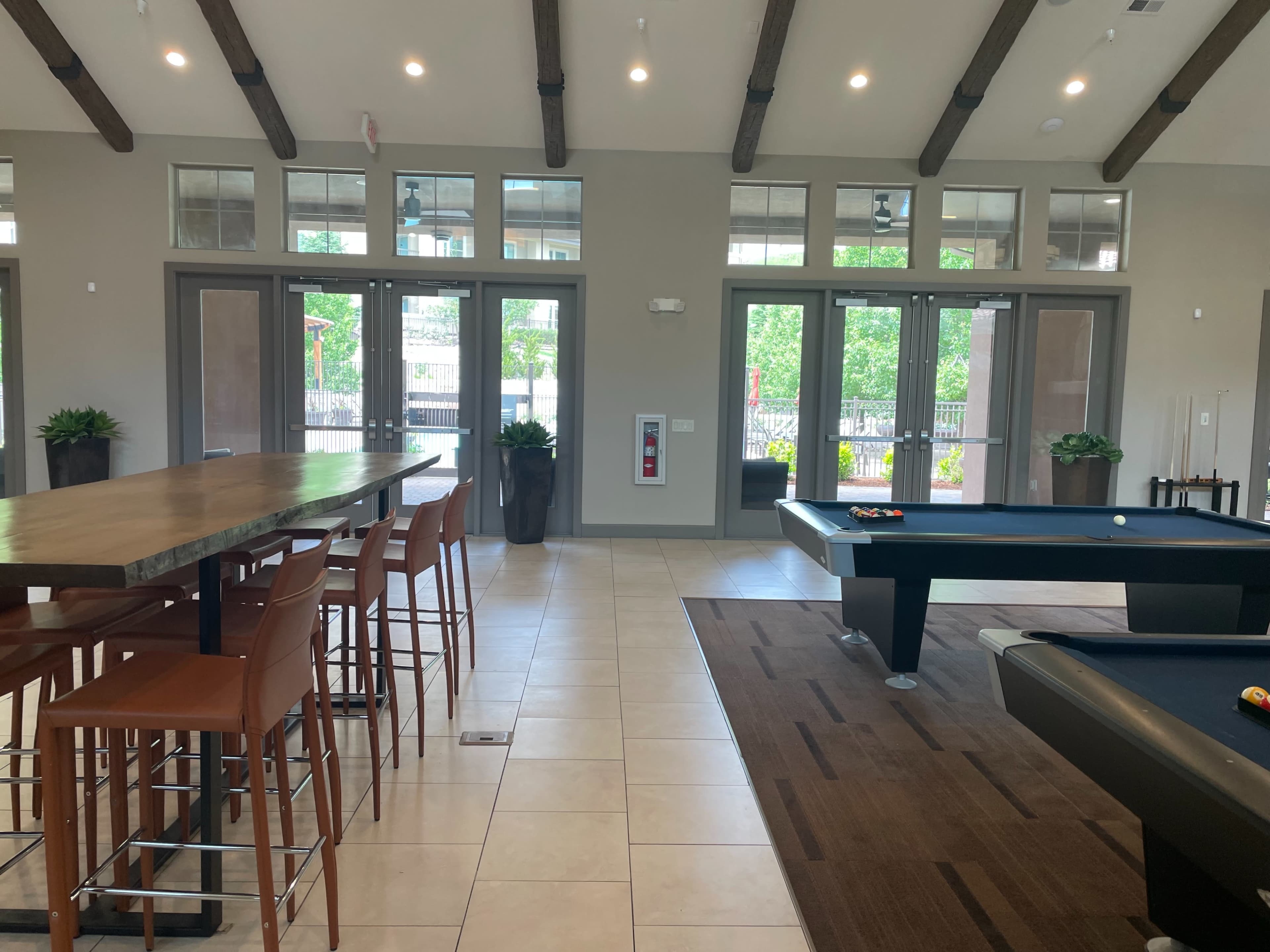 The image shows a spacious interior with a pool table, seating area with high chairs, and large windows letting in natural light.