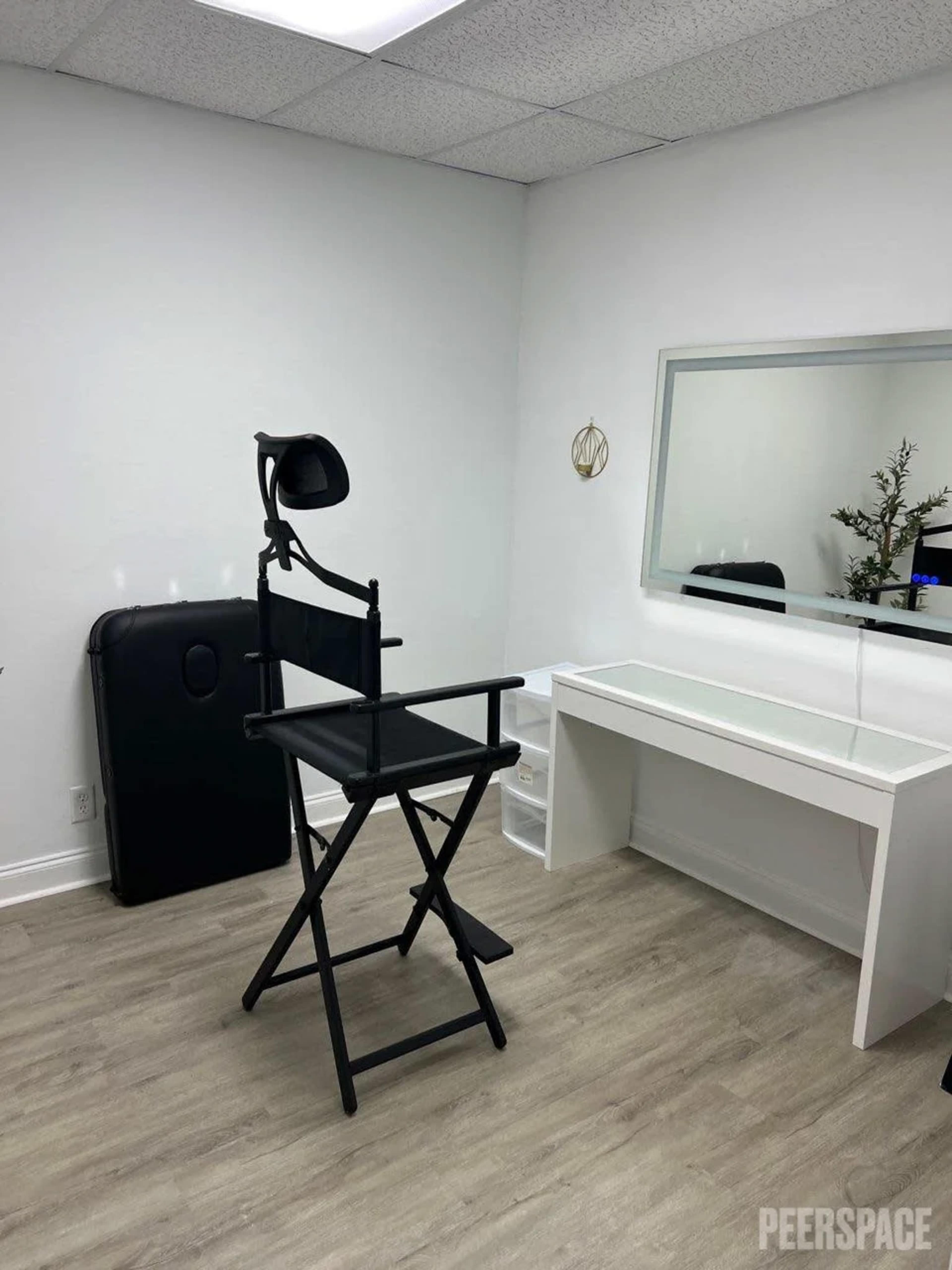 The image shows a minimalist room with a black director's chair, a white vanity table, a large mirror, and a black portable backdrop.