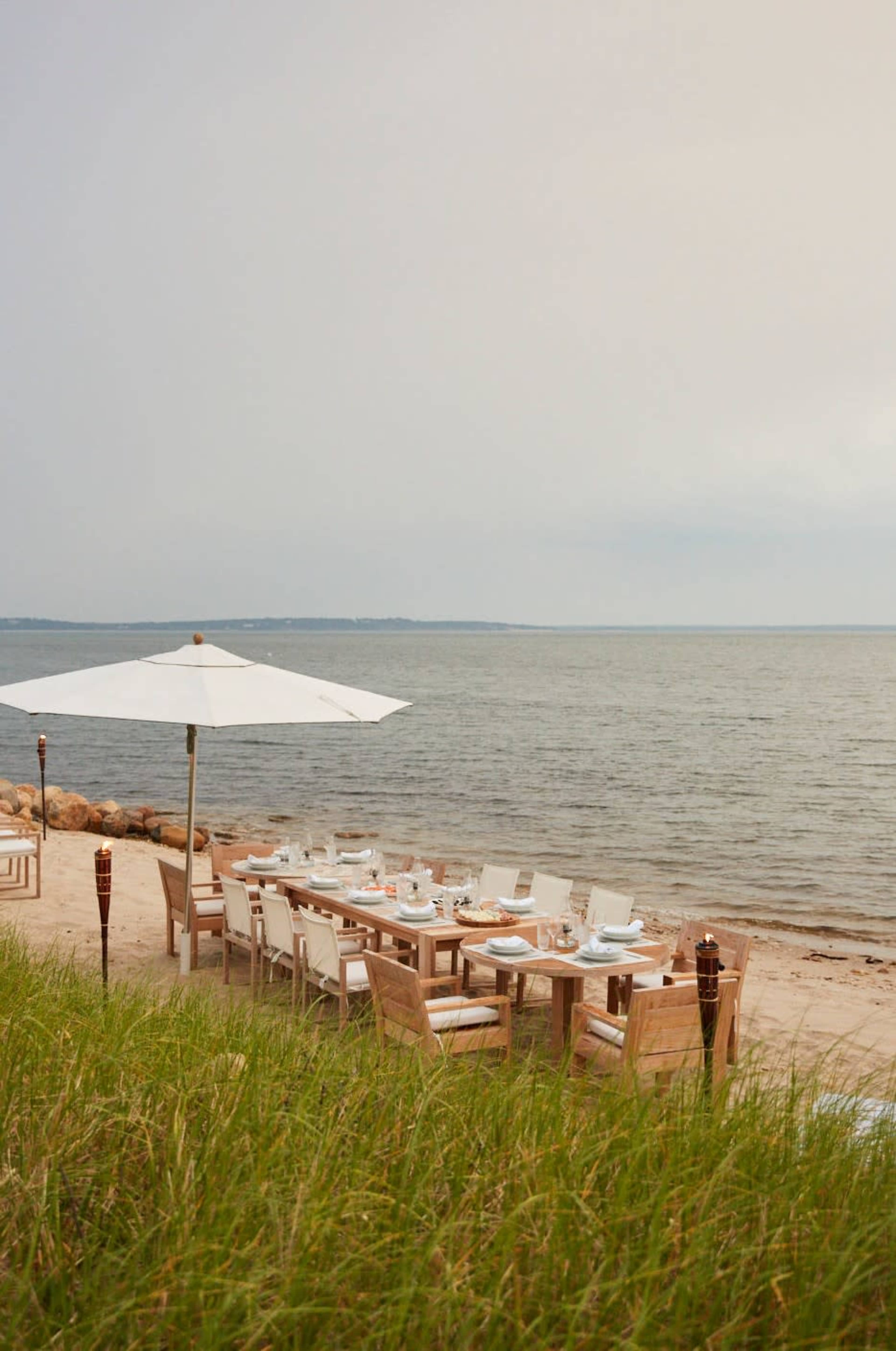 A long wooden dining table with white chairs is set up on a sandy beach near the water under a large umbrella.