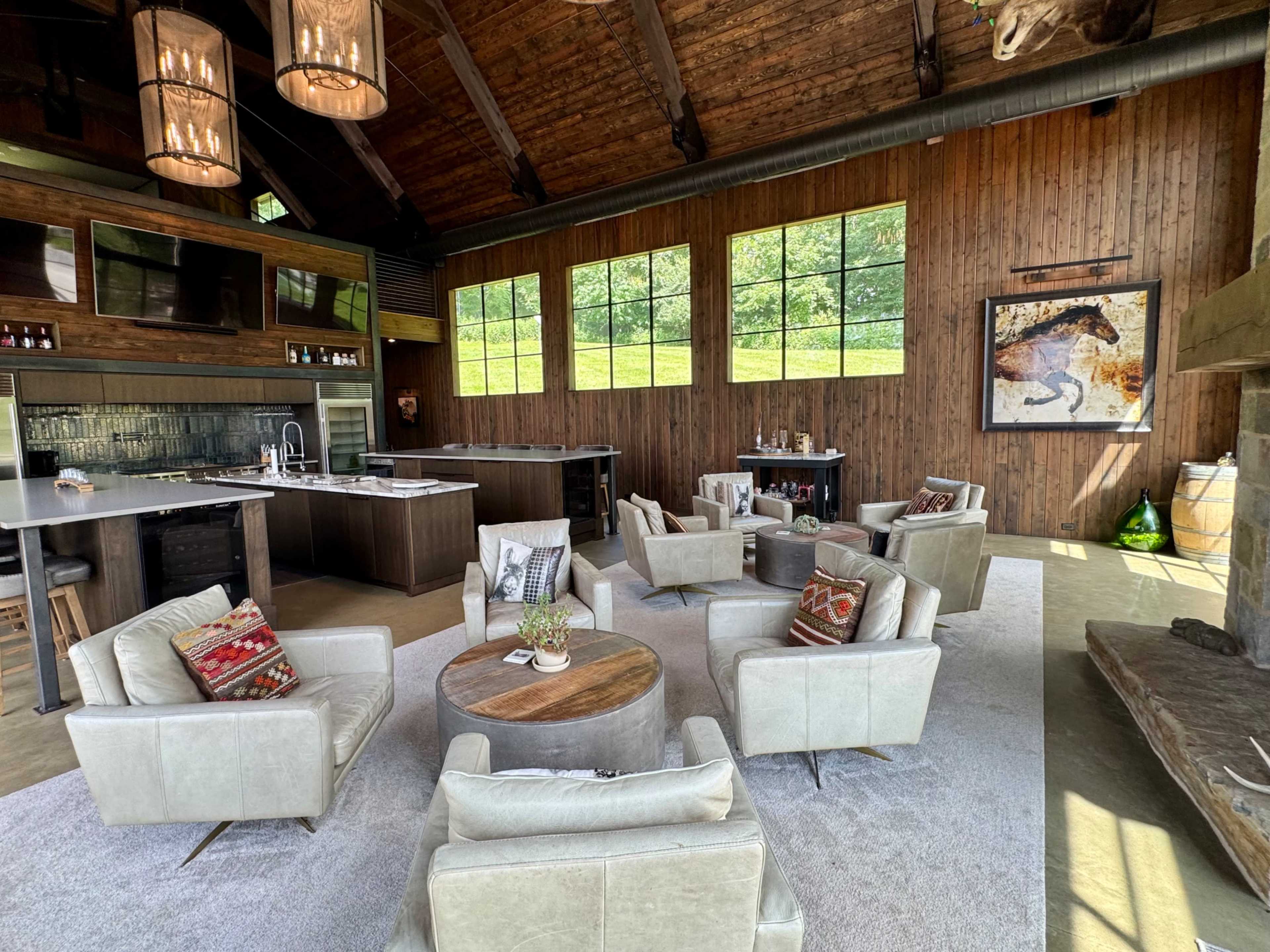 The image shows a spacious, rustic interior featuring a modern kitchen, seating area with light-colored armchairs, and large windows letting in natural light.