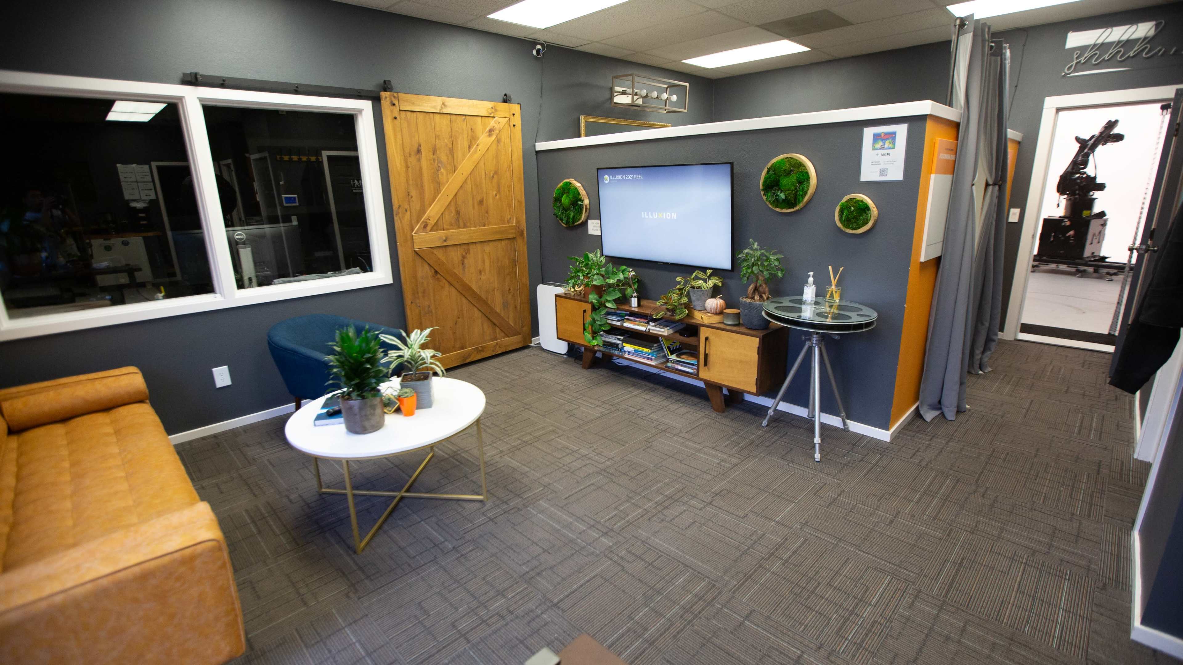 The image shows a modern office space with a cozy sitting area, a large television mounted on the wall, and decorative plants.