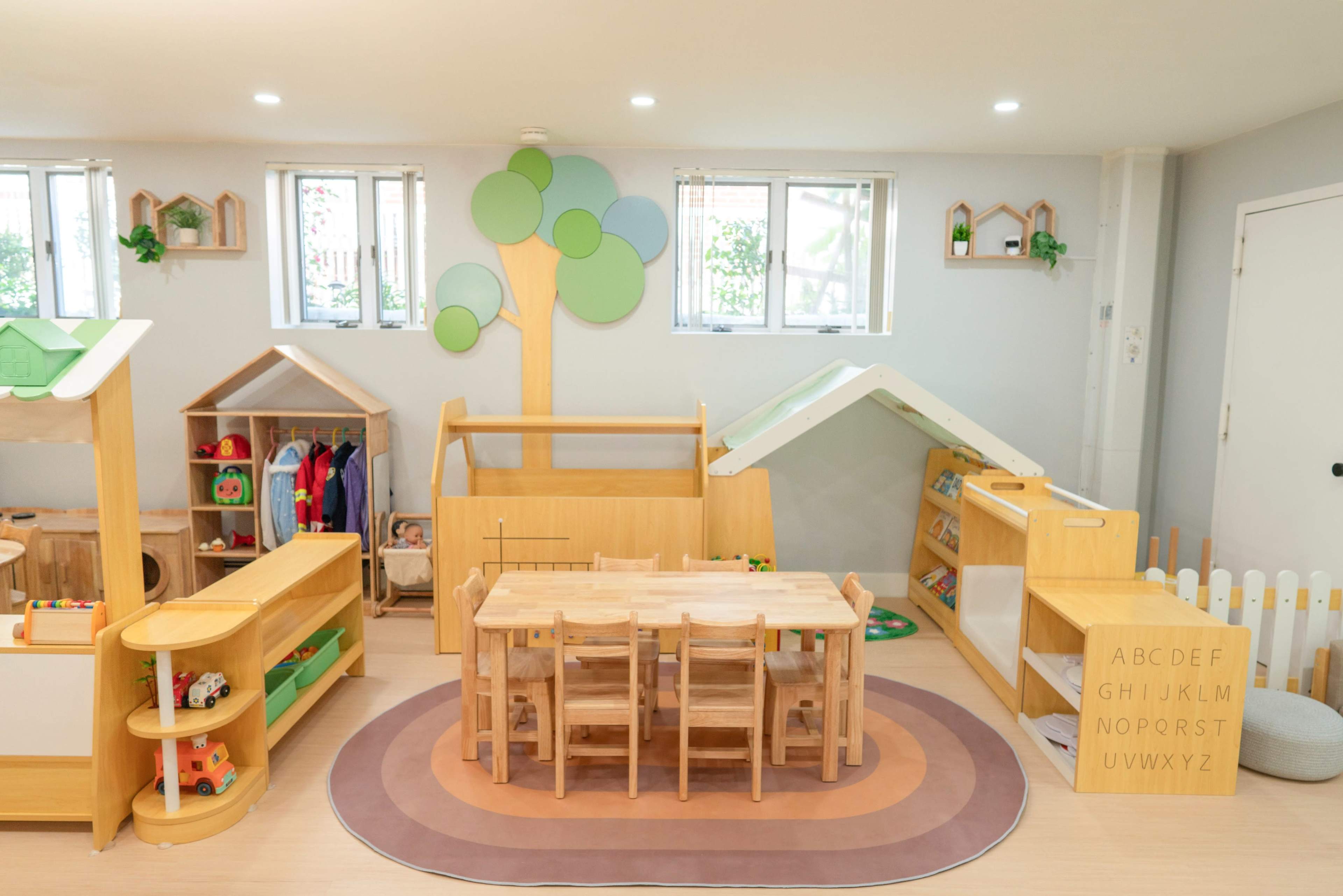 The image shows a bright indoor playroom designed for children, featuring wooden furniture, toys, and colorful decorations including a tree and playhouses.