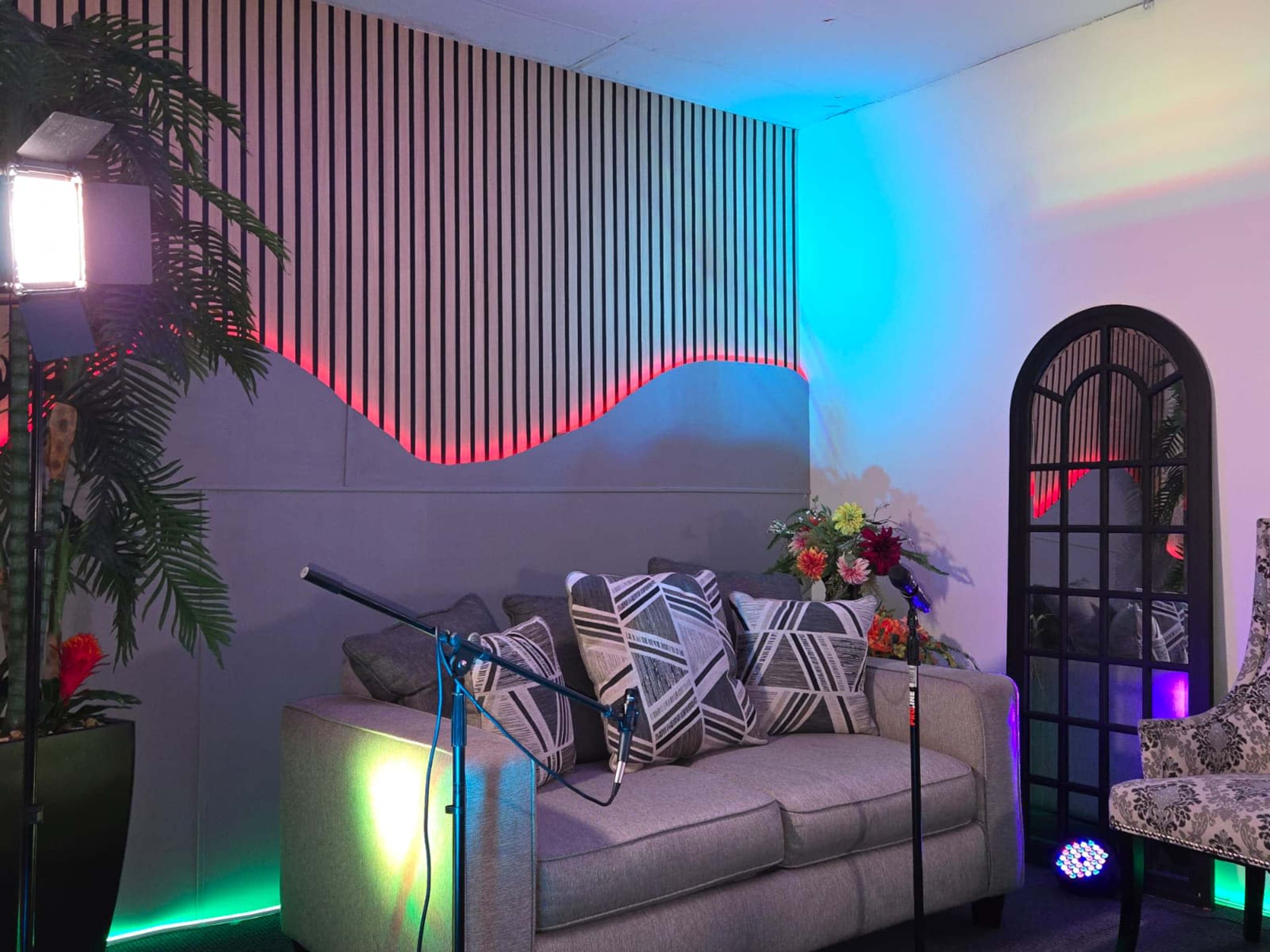 The image shows a well-decorated room with a gray sofa adorned with patterned pillows, a large mirror, and colorful lighting along the wall.
