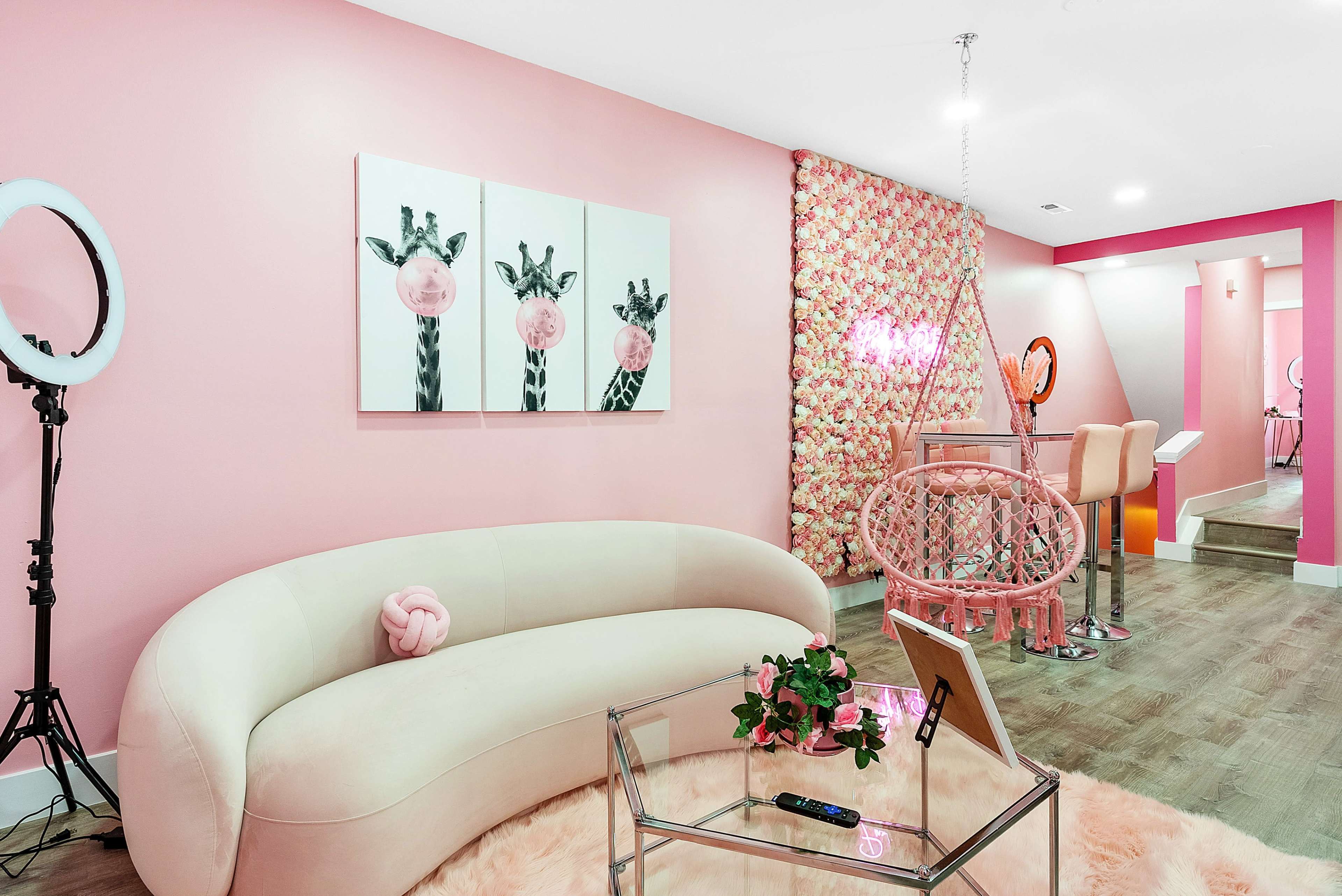 A stylish pink room featuring a curved sofa, floral artwork, a hanging chair, and a glass coffee table with a plant.