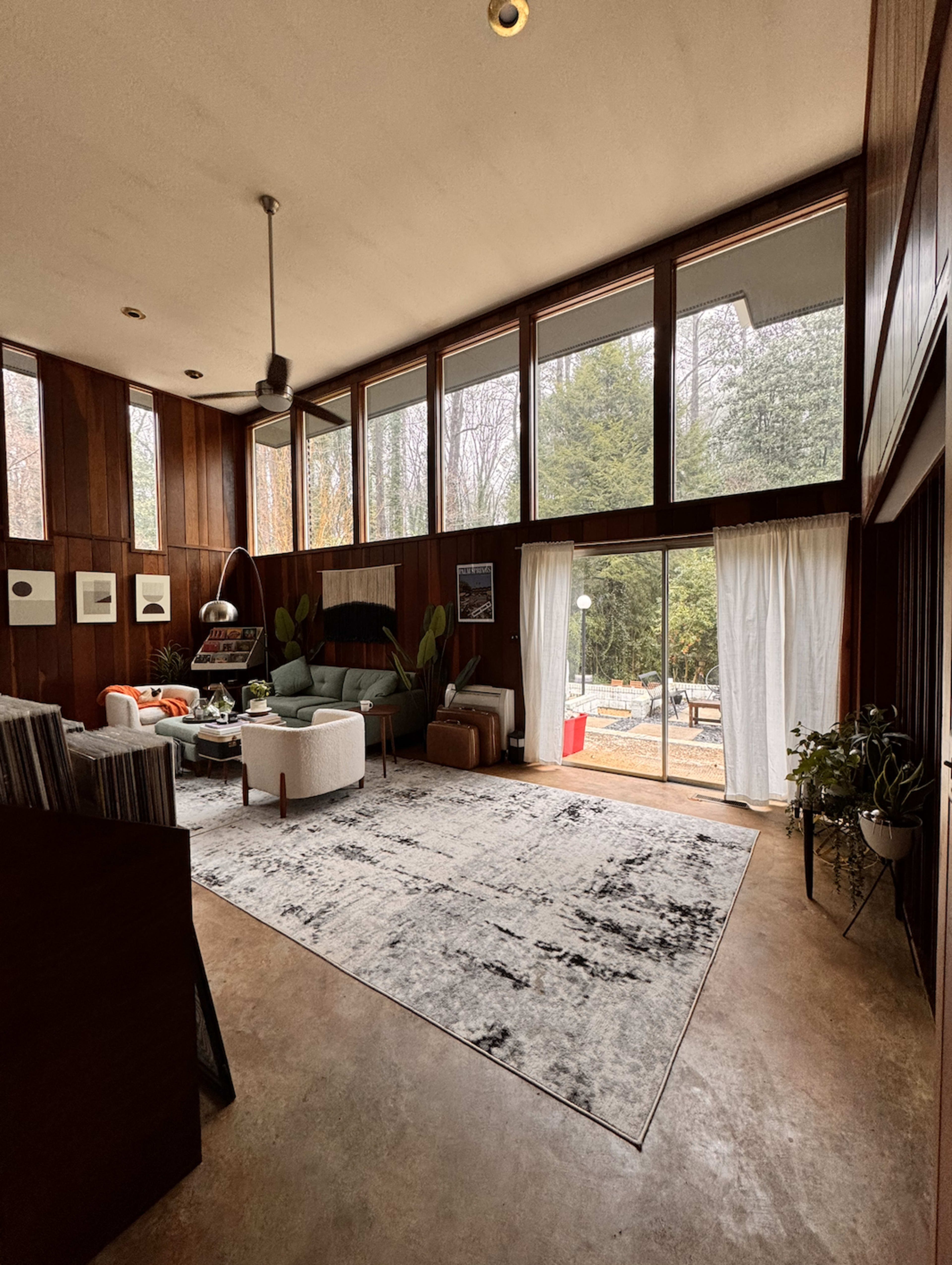 The image shows a modern living room with large windows, wooden walls, and a mix of furniture including a green sofa, white armchairs, and a round coffee table, overlooking an outdoor area.