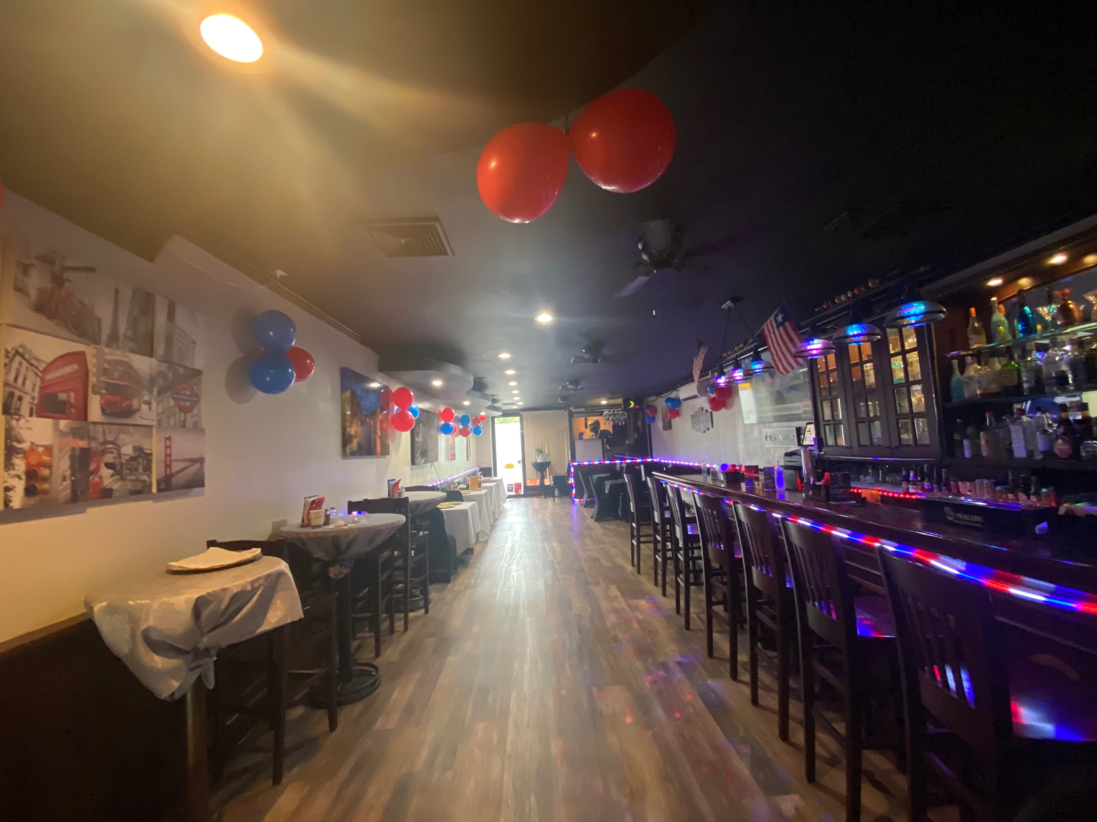 The interior of a bar is decorated with blue and red balloons, and colorful lights line the counter.