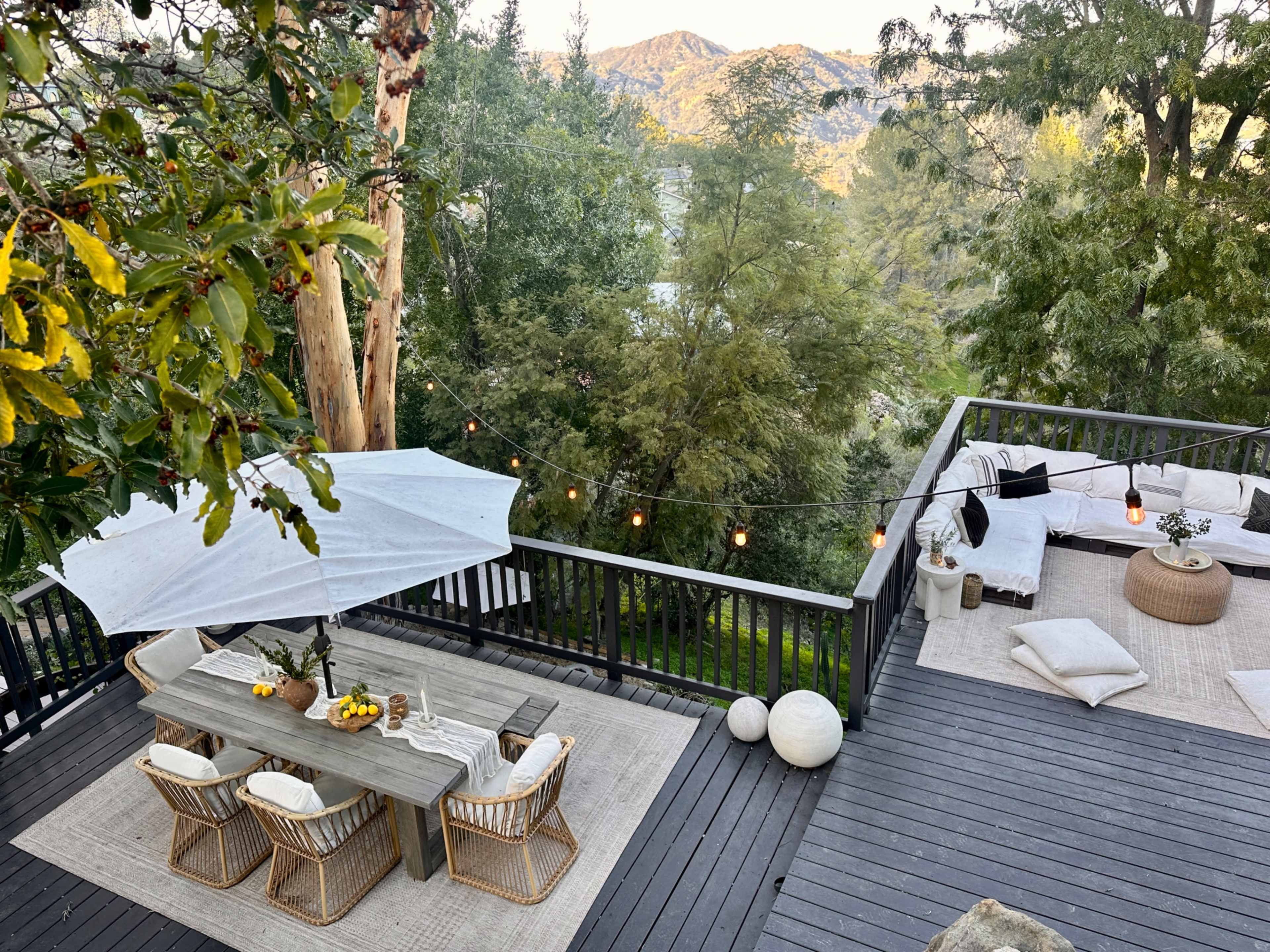 A wooden deck features a dining table with chairs, surrounded by greenery and mountain views, illuminated by string lights.