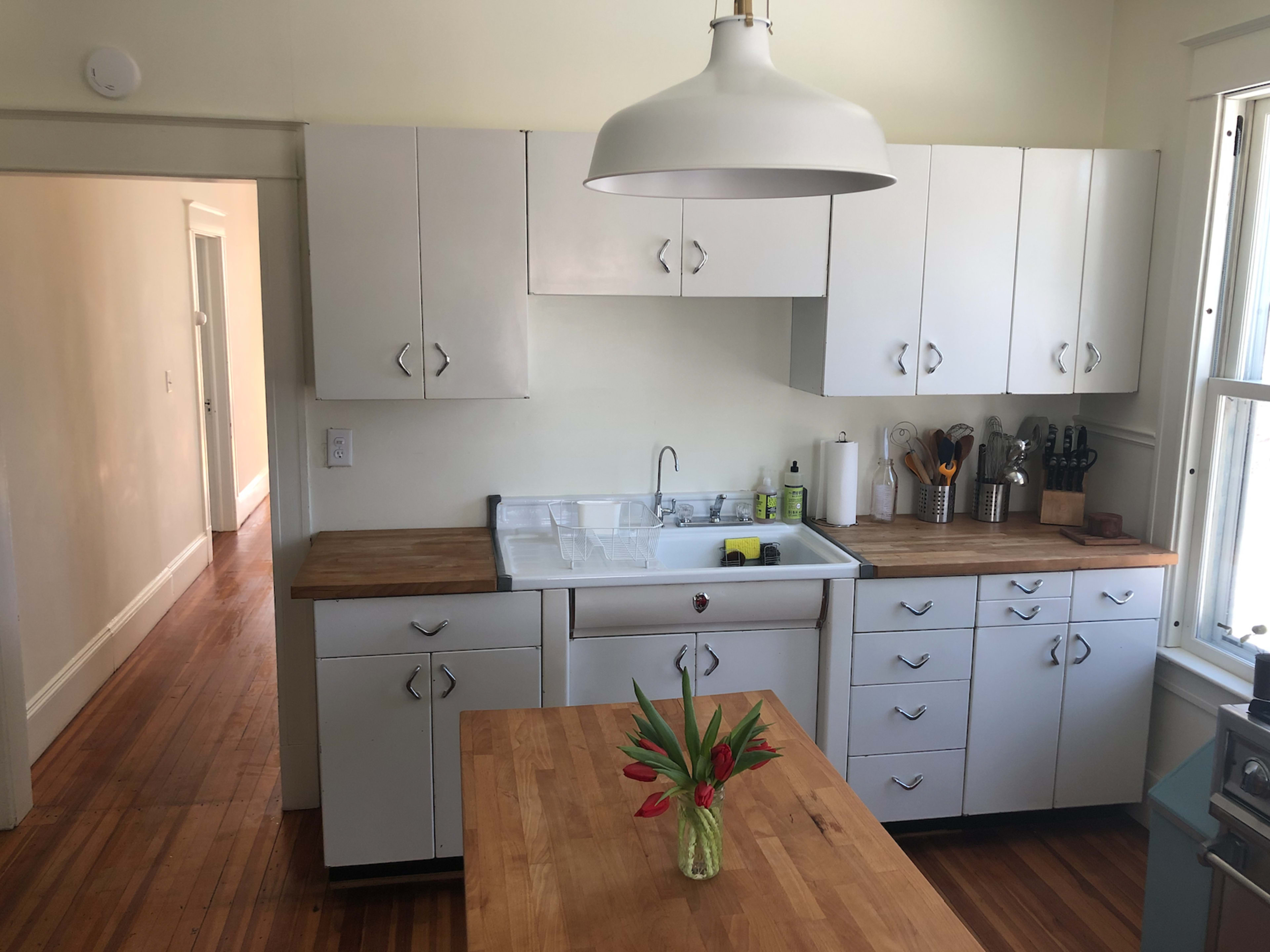 A clean kitchen features white cabinets, a wooden countertop, and a sink with a window above it, complemented by a vase of flowers on the table.