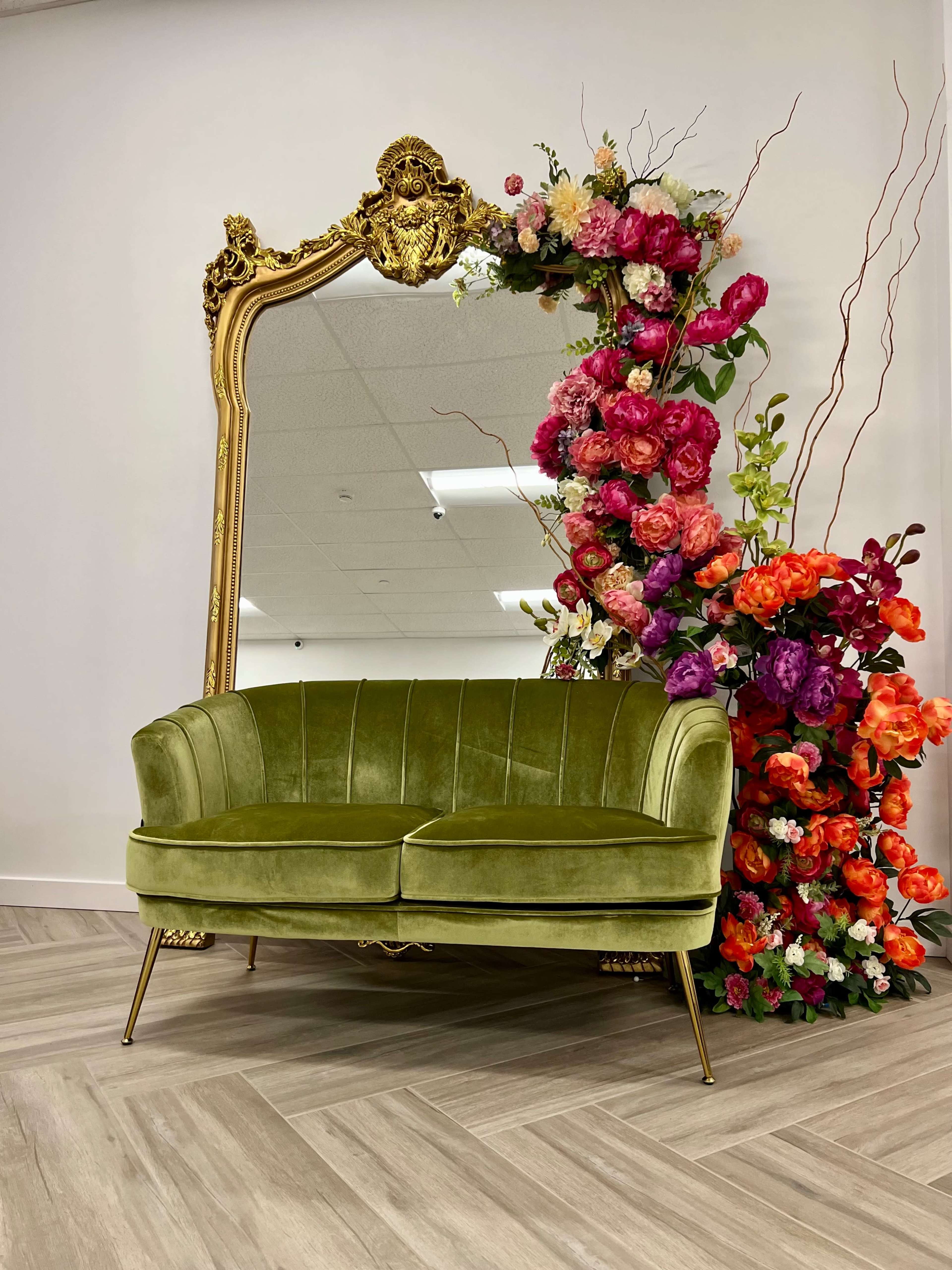 A green velvet sofa is positioned in front of a large ornate mirror framed with a colorful arrangement of flowers.
