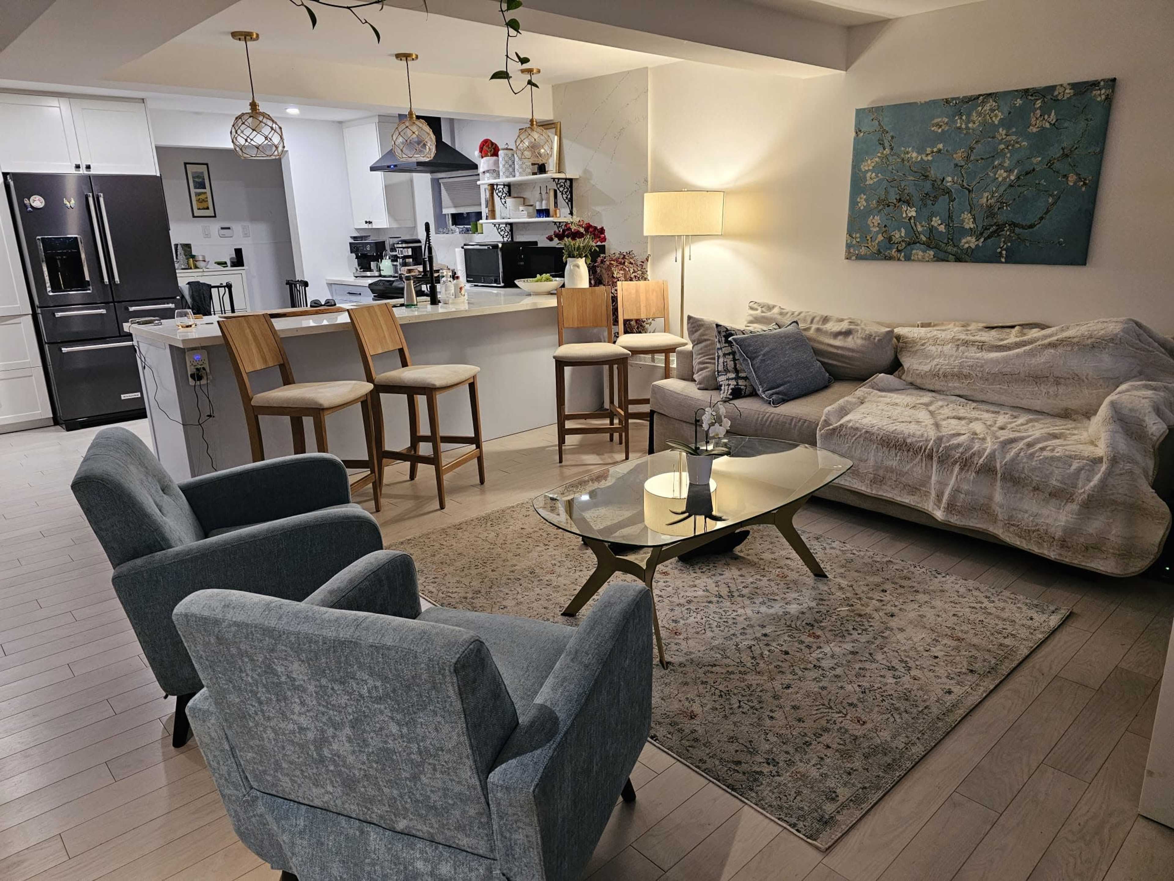 A cozy living room features a sofa, two armchairs, a coffee table, and a kitchen bar area with stools in the background.