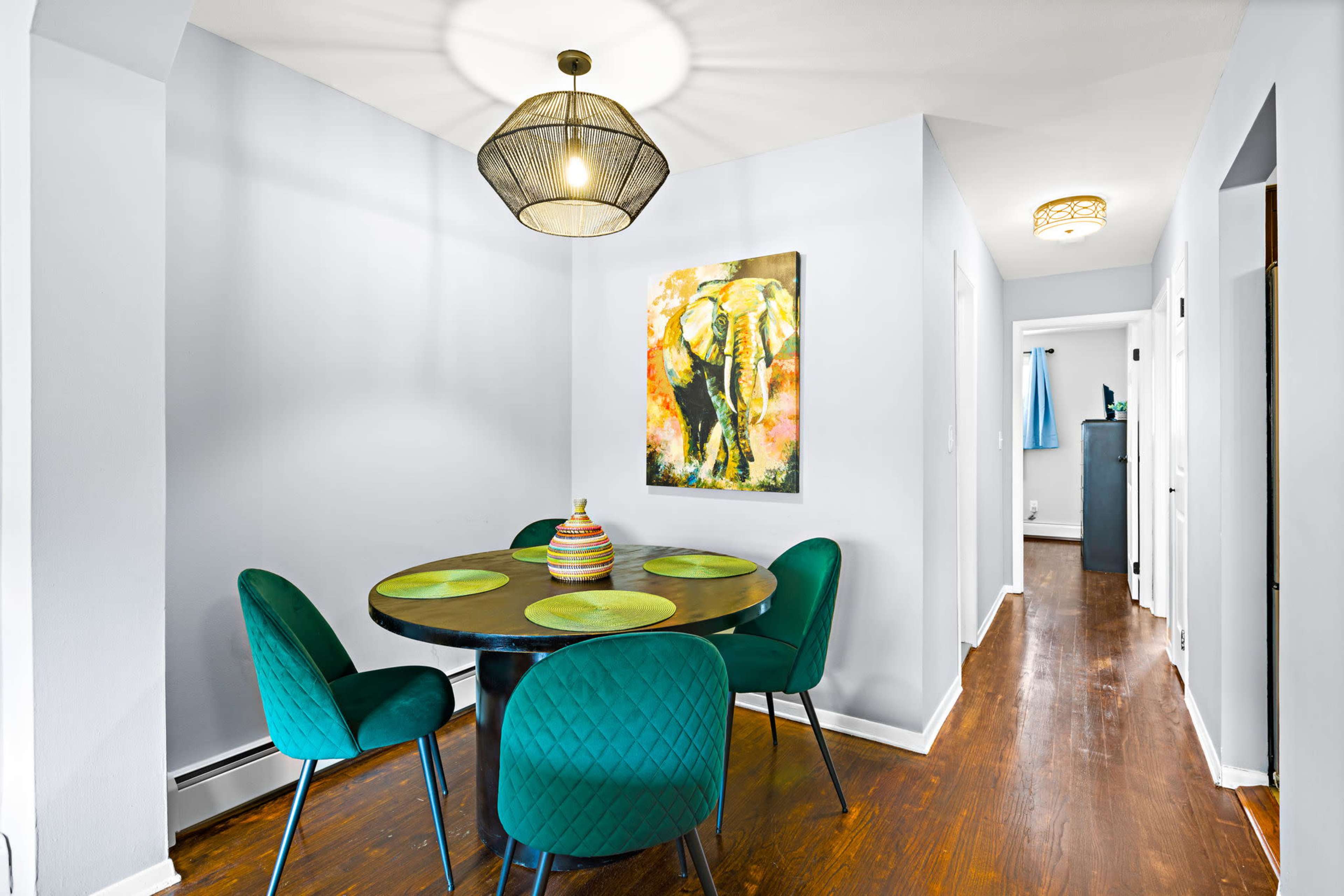 A dining area features a round table with green place mats surrounded by teal chairs, illuminated by a pendant light, and a colorful elephant painting is hung on the wall.