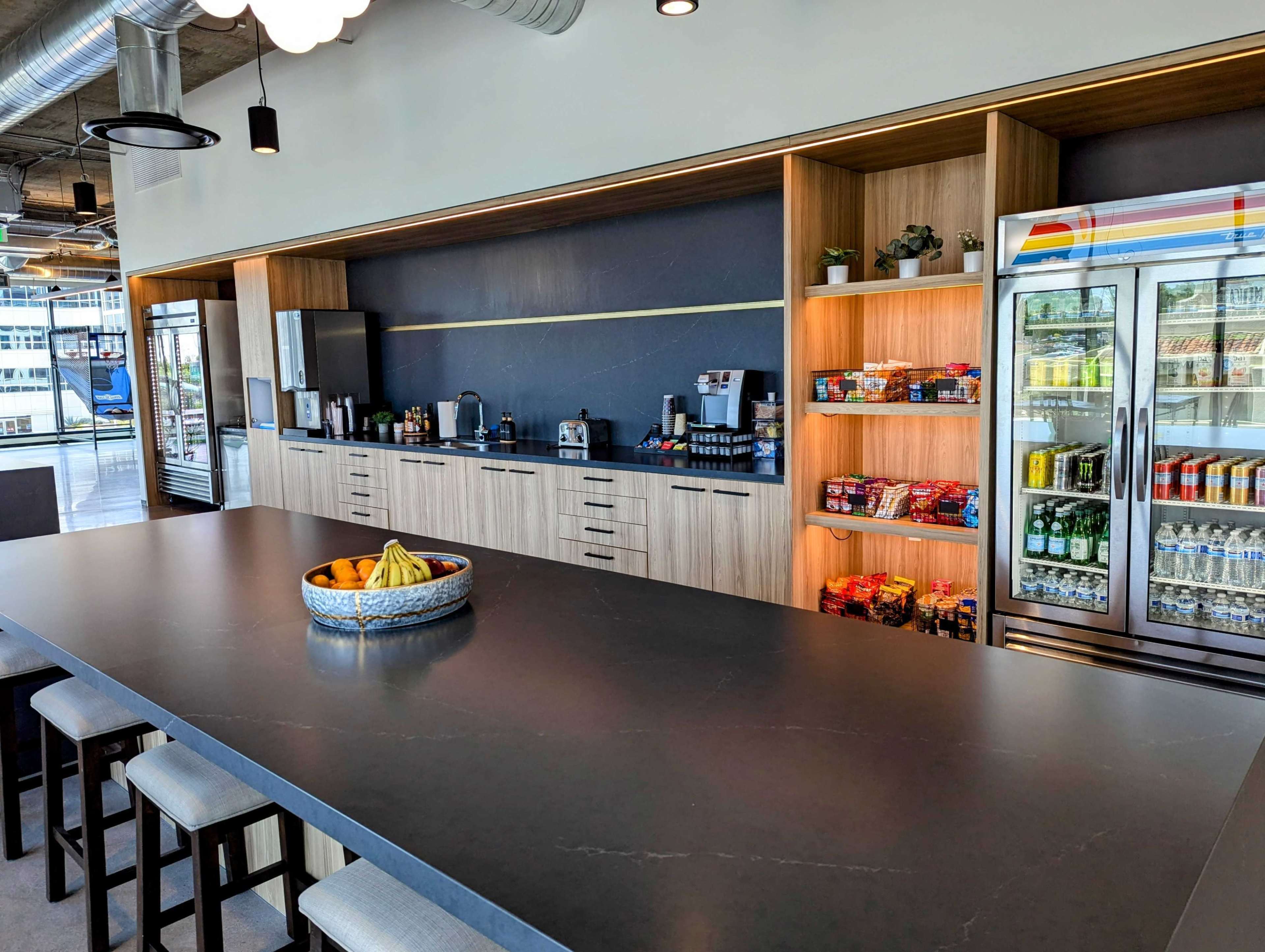 A modern kitchen area with a large dark countertop, shelves of snacks, beverages in a glass fridge, and various kitchen appliances against a wooden backdrop.