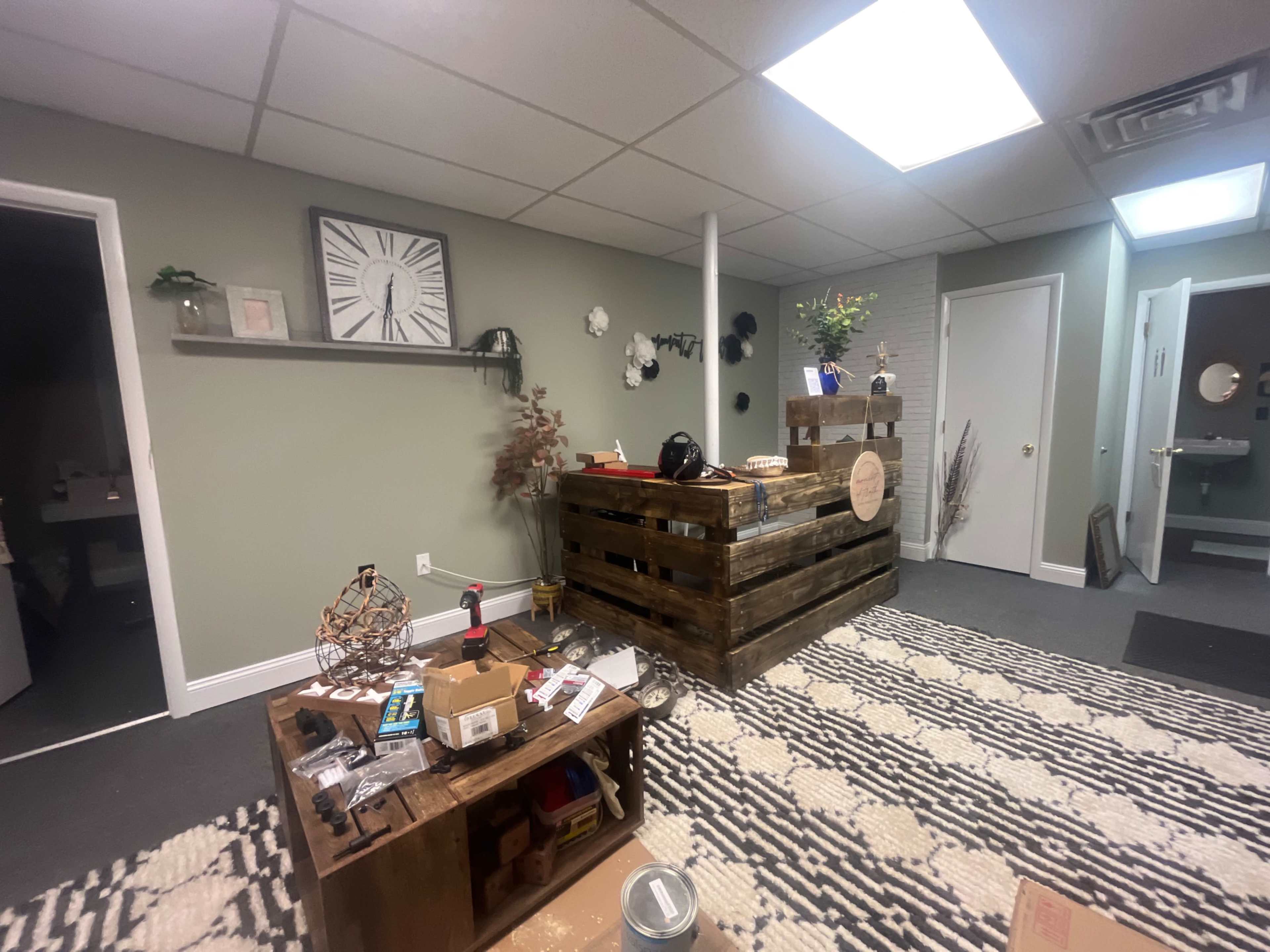 The image shows a modern workspace featuring a reception area with a wooden counter made from pallets, a small decorative table with scattered items, and a door leading to a restroom in the background.