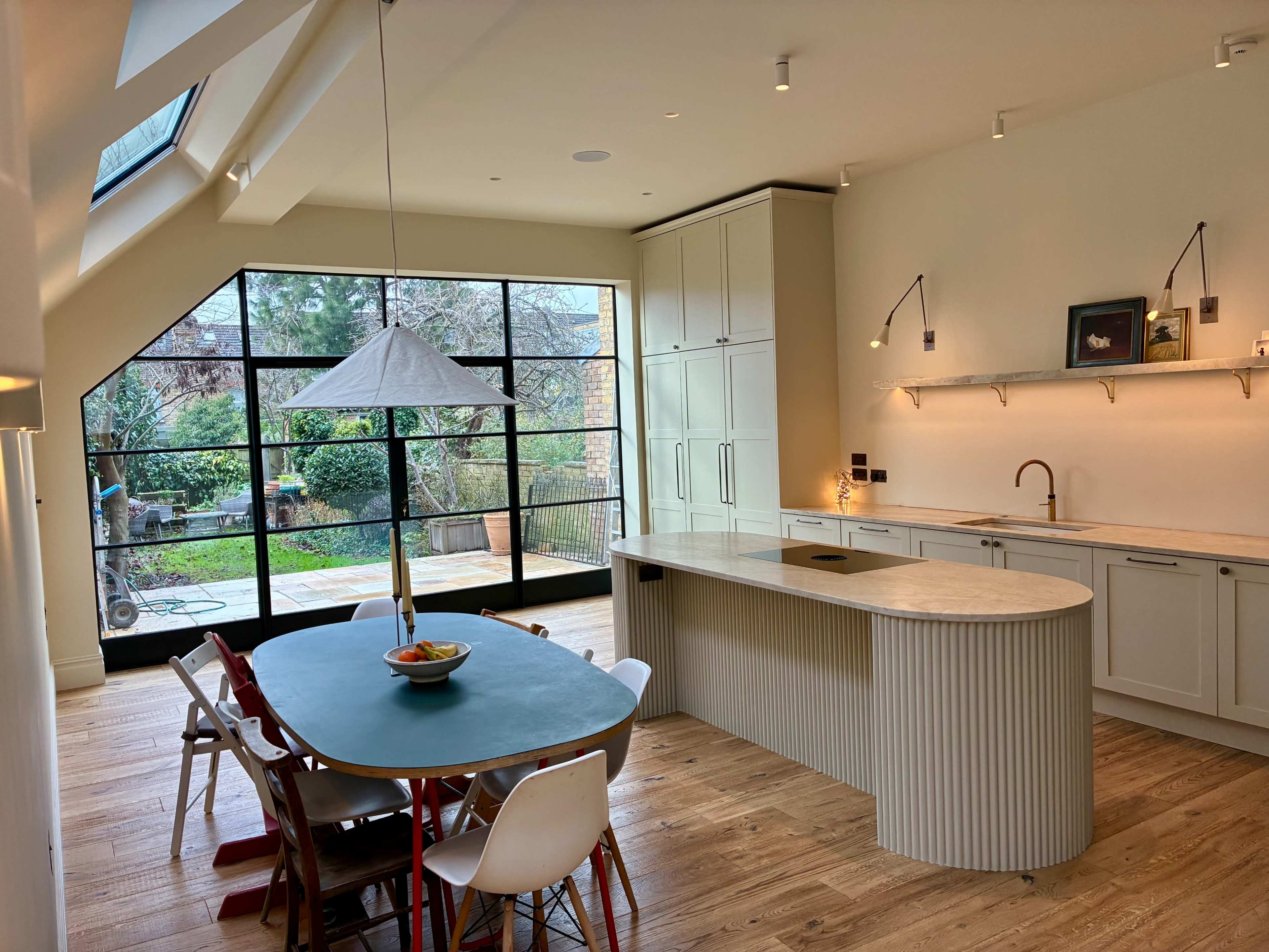 The image shows a modern kitchen with an island counter, a dining table, and large windows overlooking a garden.