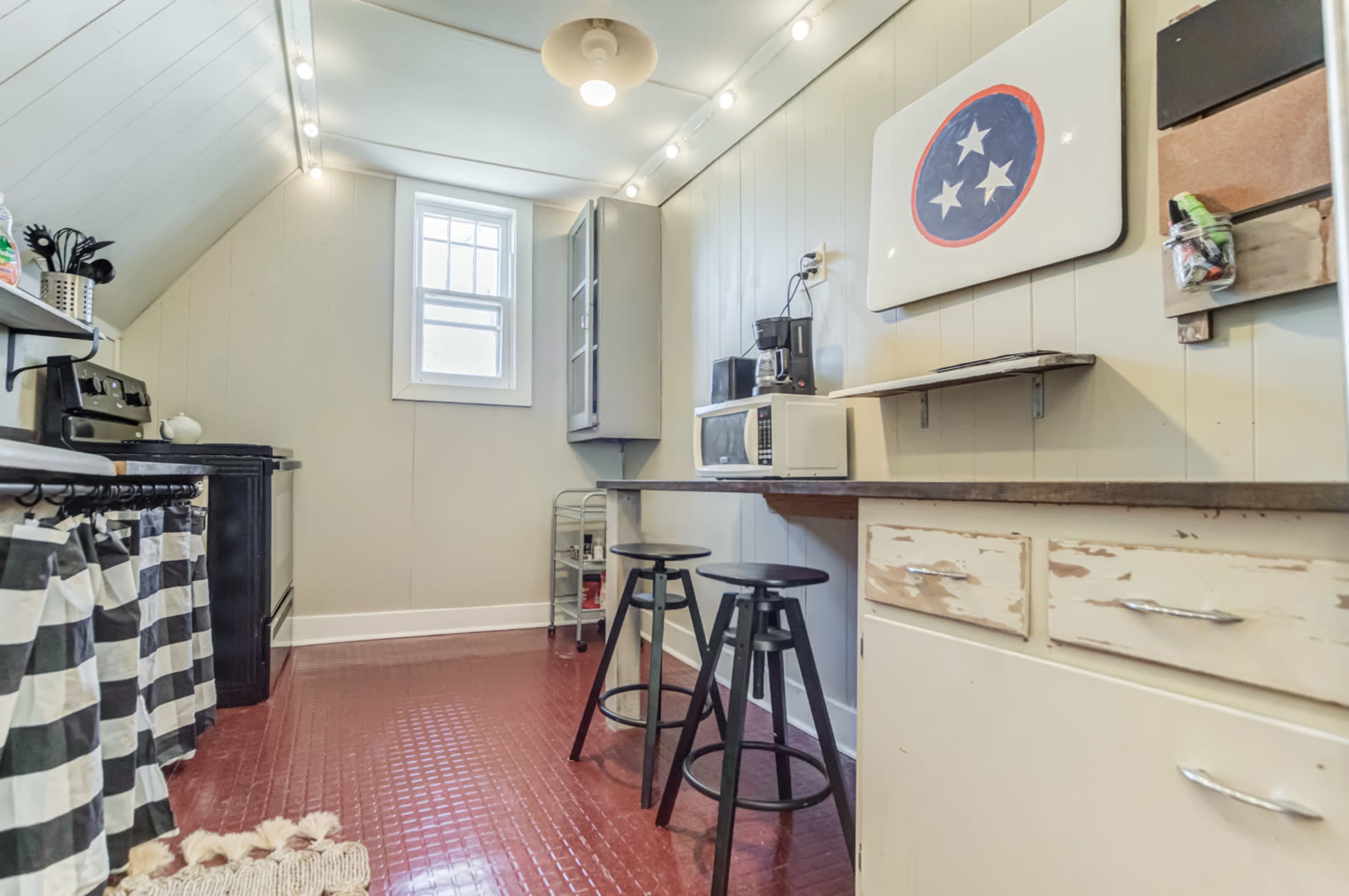 A compact kitchen with a black stove, microwave, and two black stools next to a countertop, featuring light-colored walls and checkered curtains.