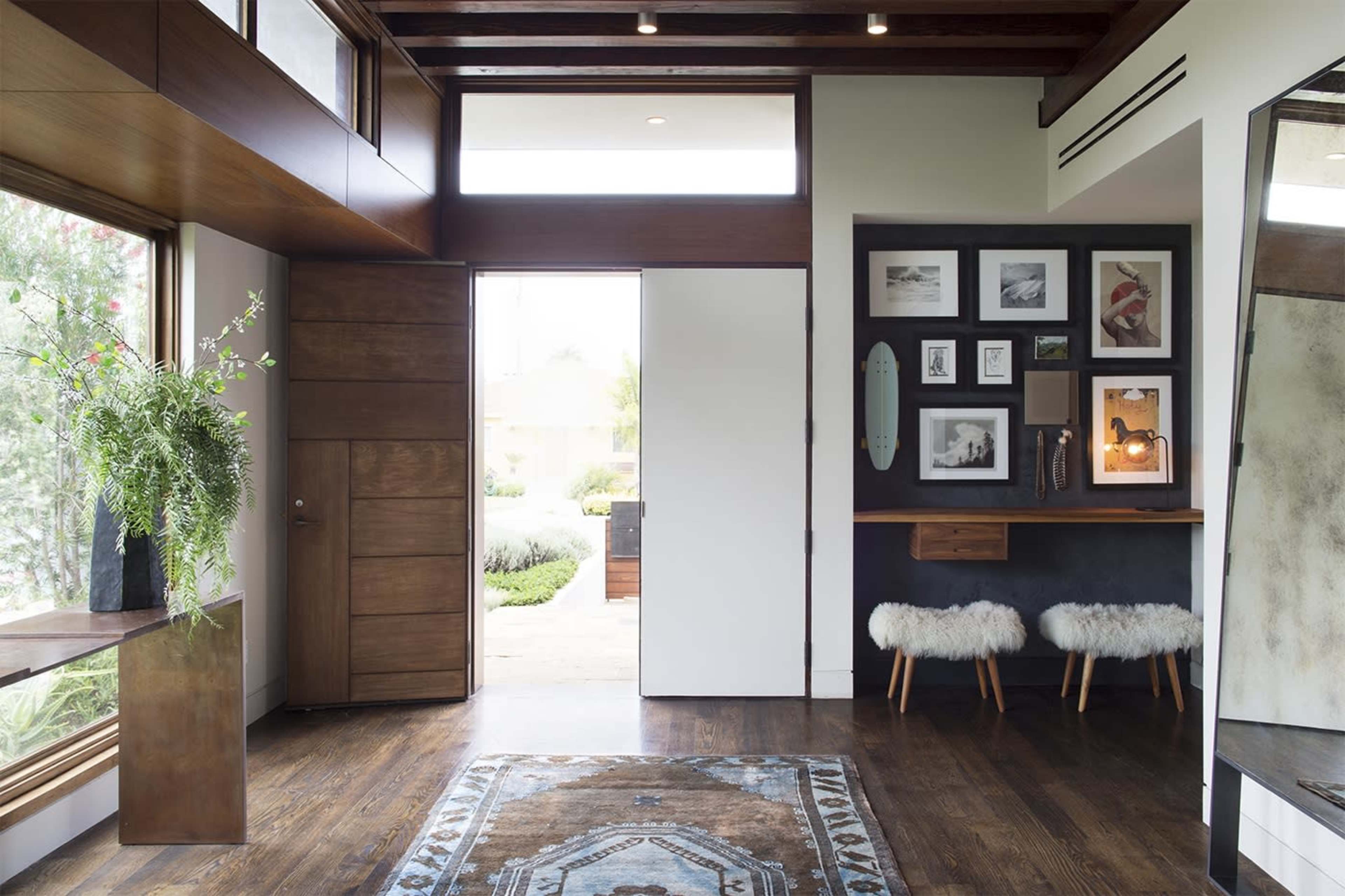 A modern entryway featuring a large window, a door leading outside, a small table with a decorative arrangement, and a wall adorned with framed pictures next to two upholstered stools.
