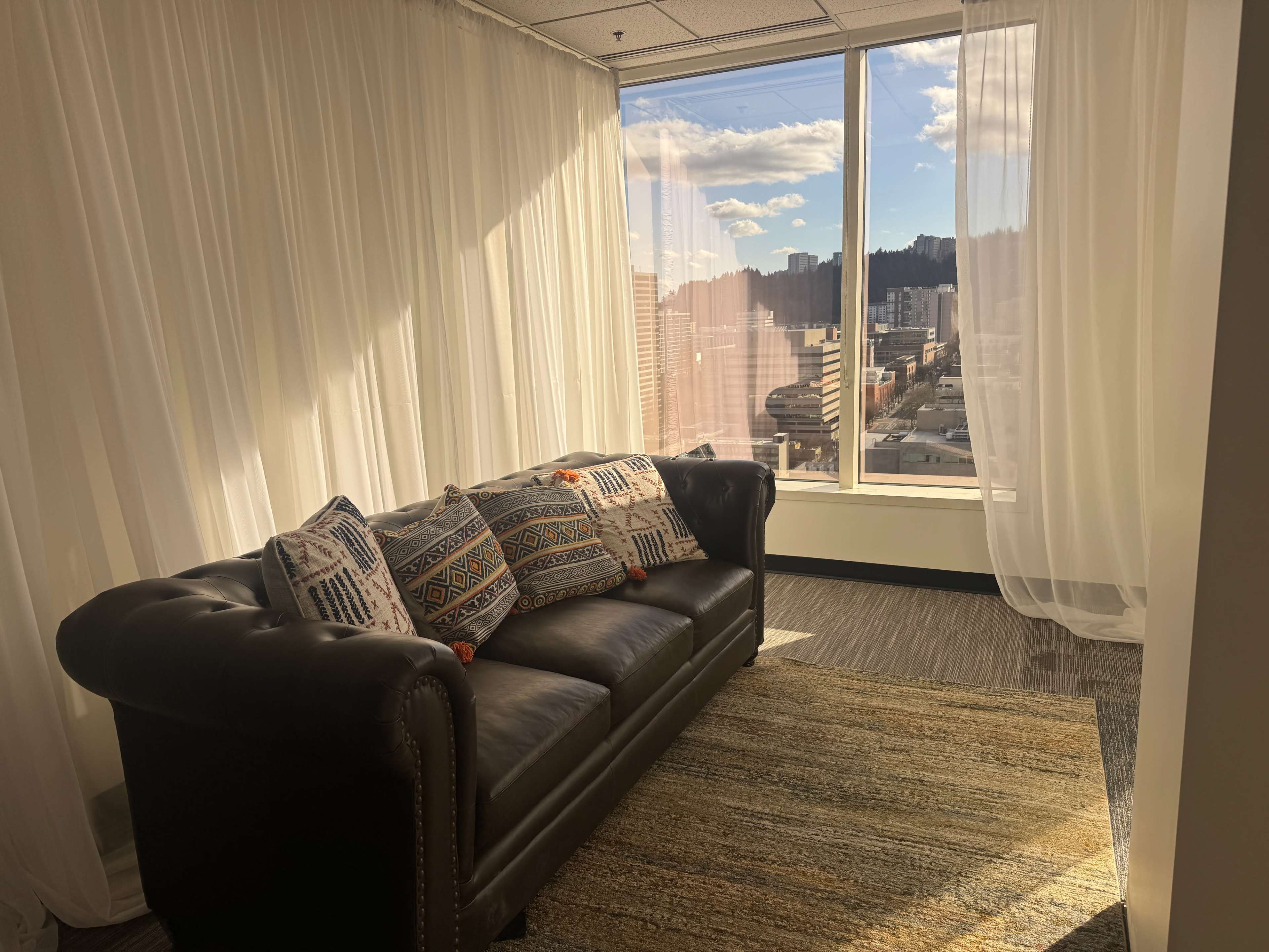 A leather sofa with decorative pillows sits beside a large window, allowing natural light to illuminate the space and offering a view of the city outside.