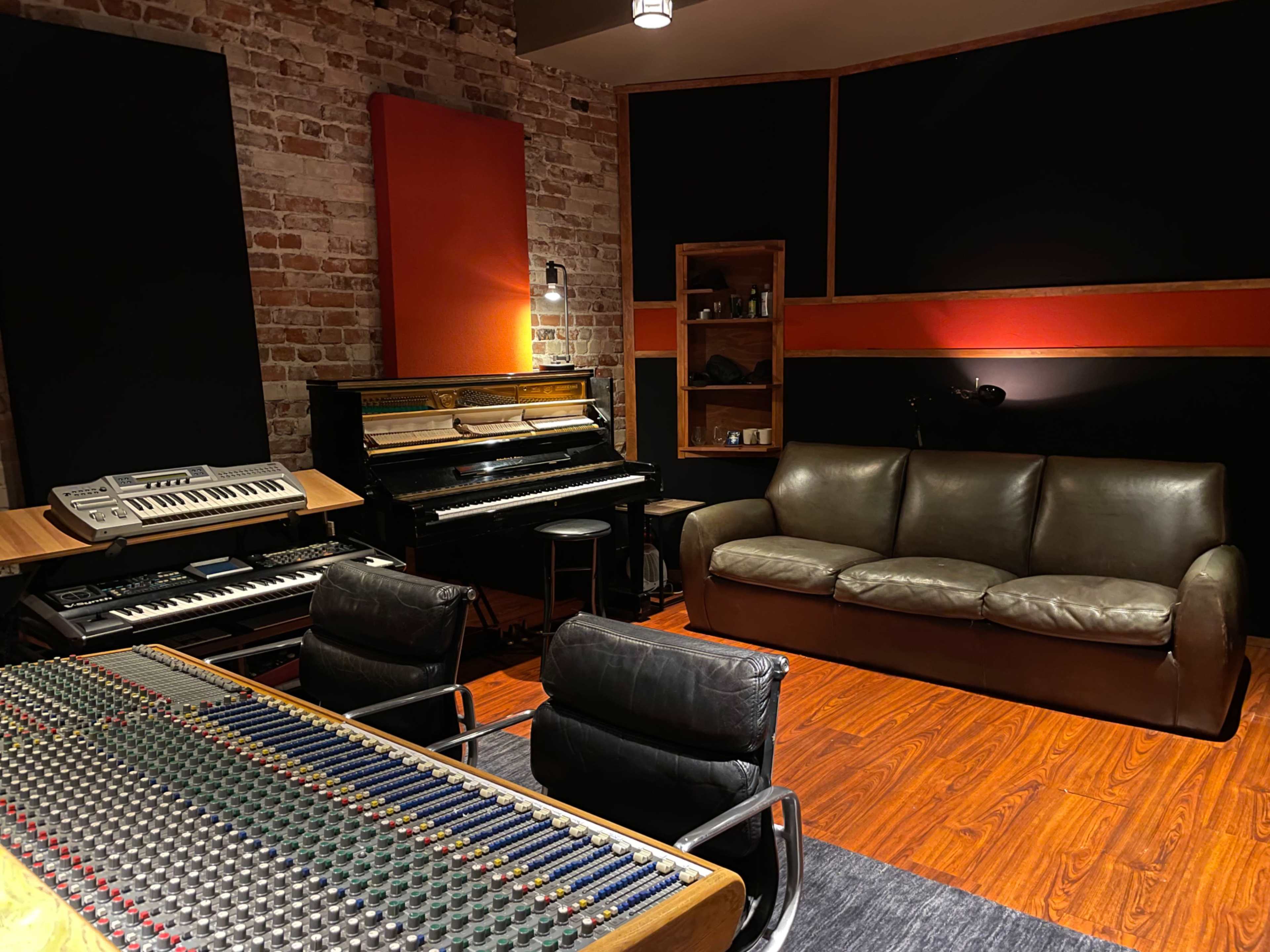 A music studio featuring a mixing console, a piano, keyboards, a sofa, and exposed brick walls.