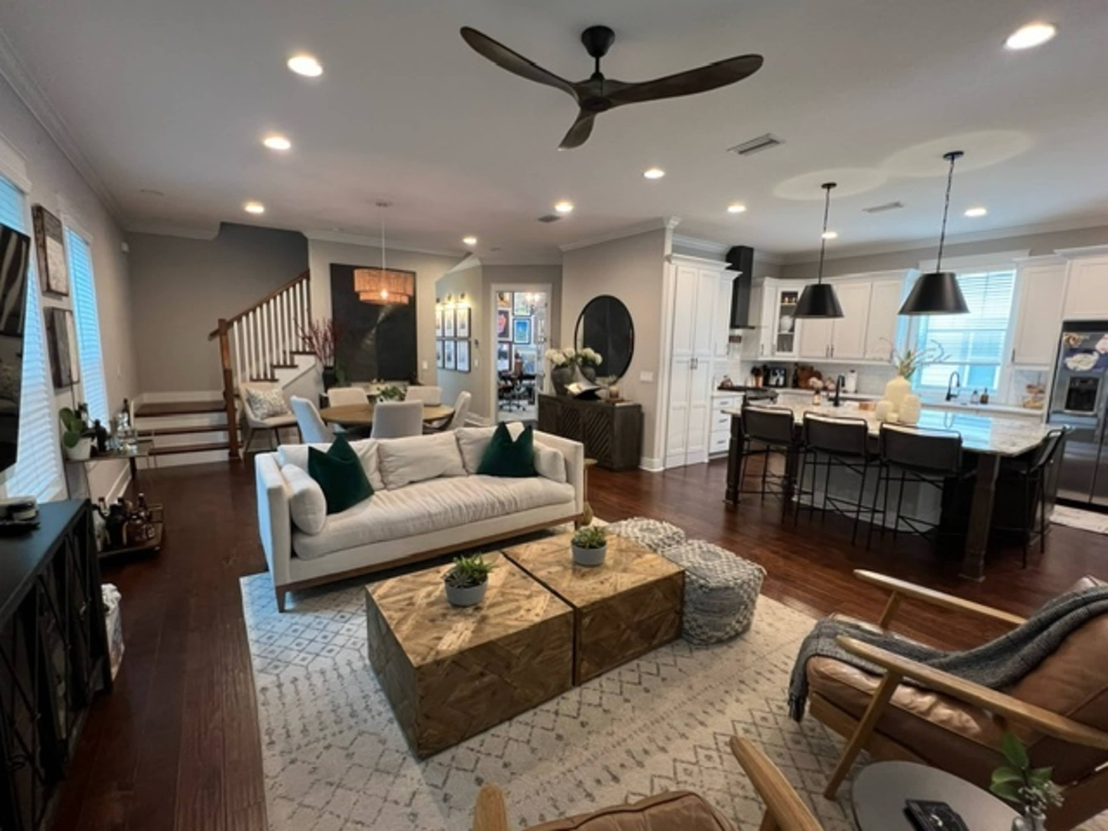 A modern living room with a sectional sofa, two wooden coffee tables, and a kitchen in the background featuring white cabinetry and bar seating.