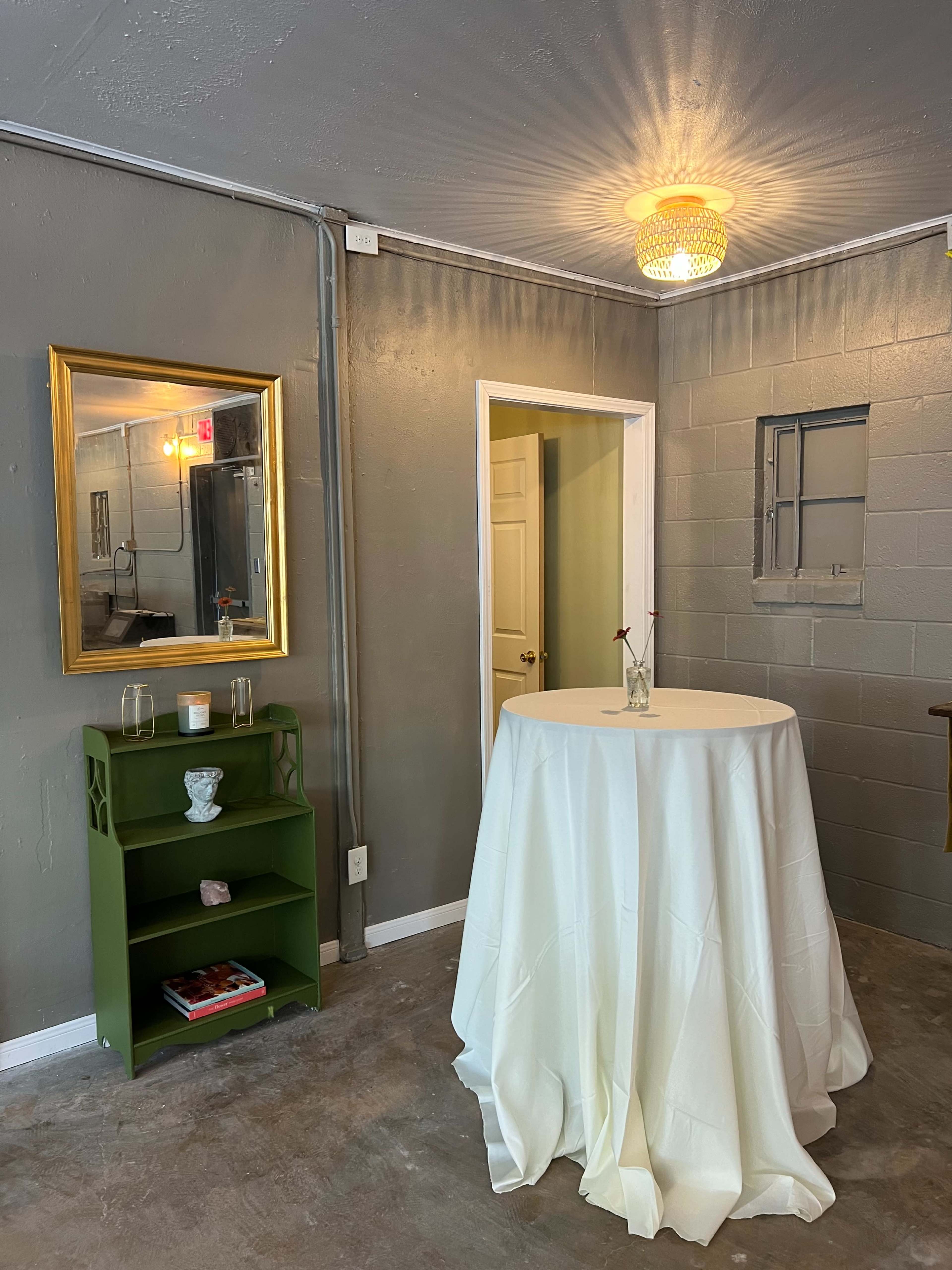 The image shows a small room with gray walls, a round table covered with a white tablecloth, and a green shelf holding decorative items, illuminated by a pendant light.