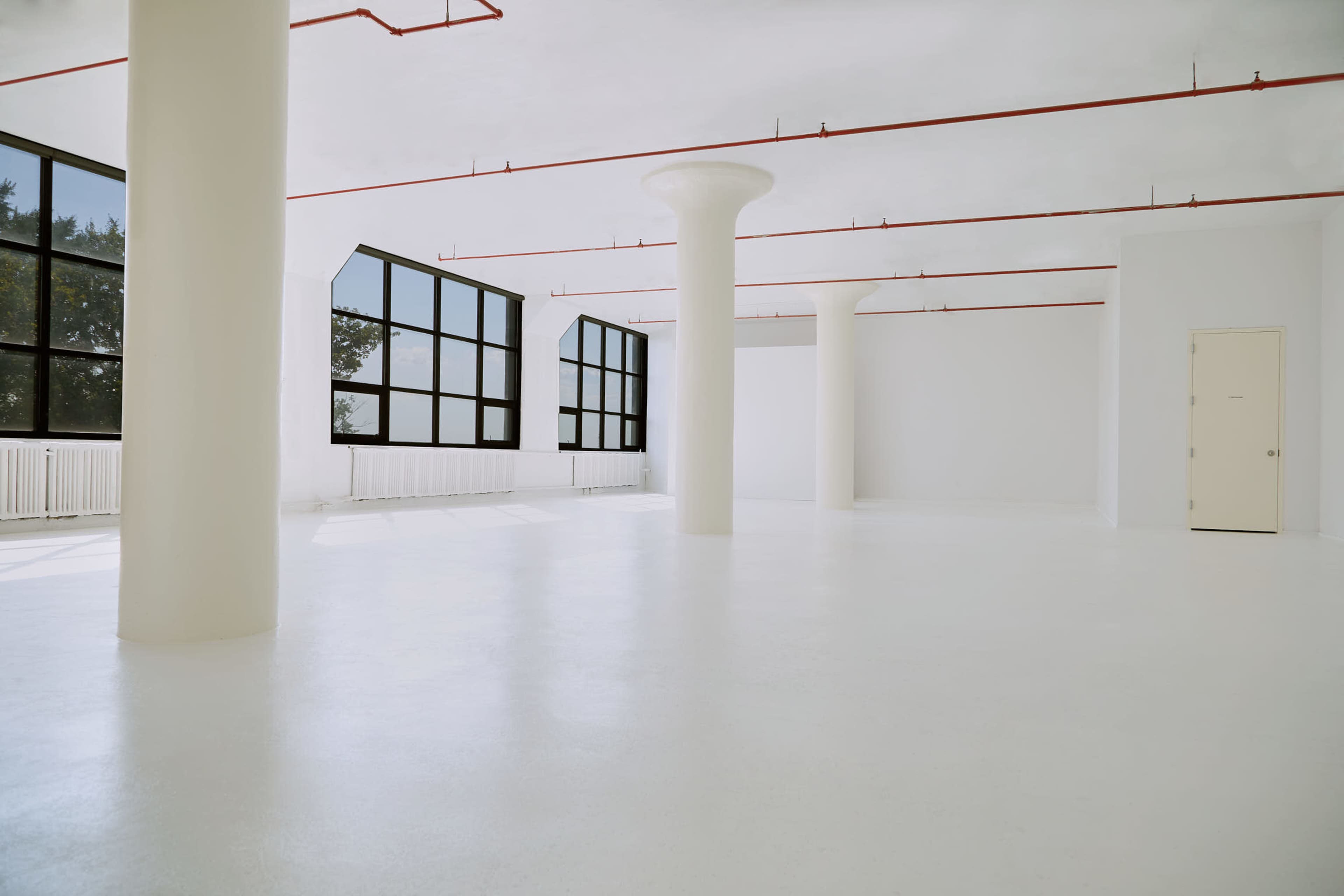 A spacious, empty room with white walls, a smooth white floor, large windows, and several white columns.