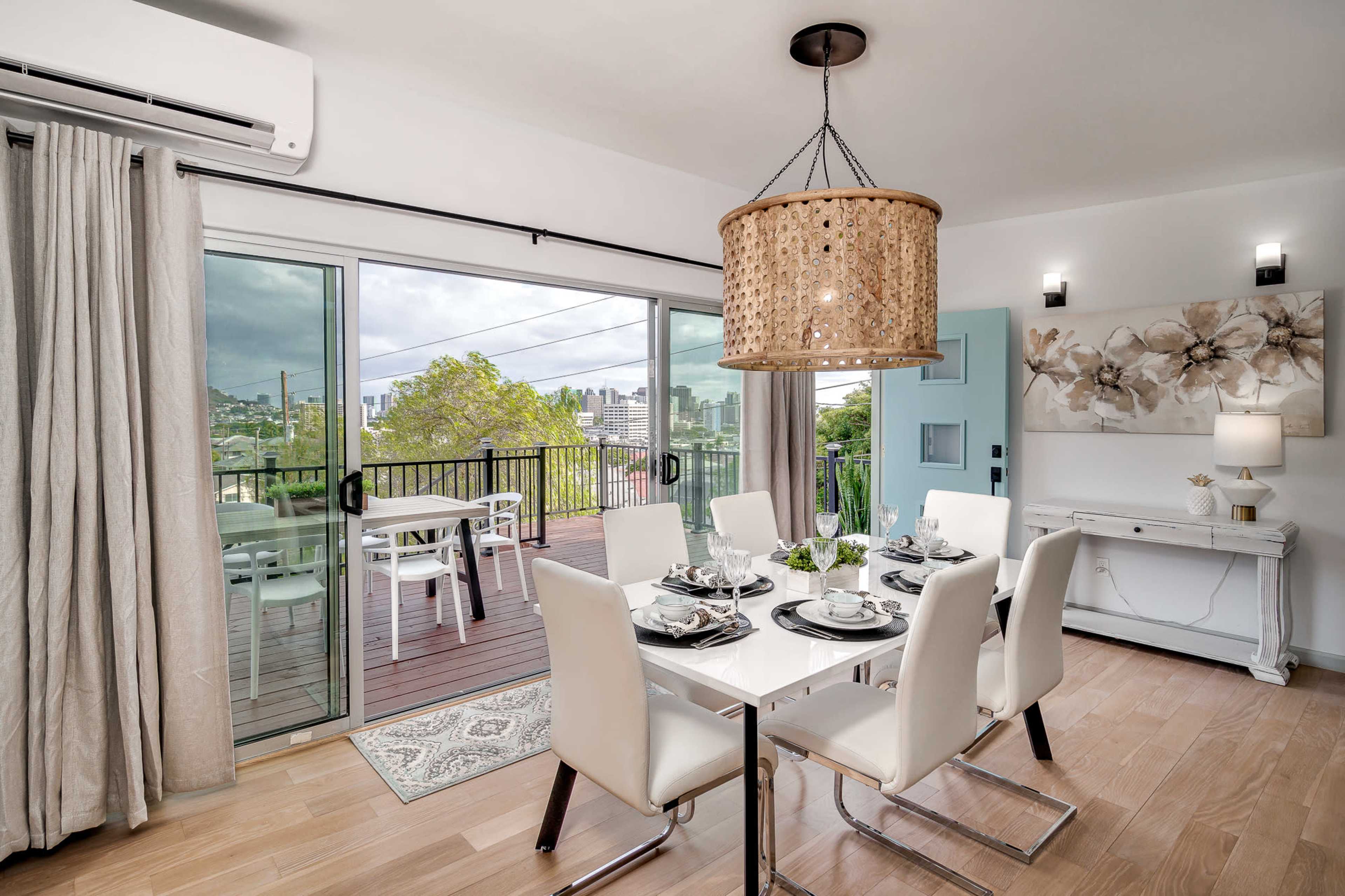 A dining room features a white table set for six, with large windows offering a view of the city and a door leading to a balcony.