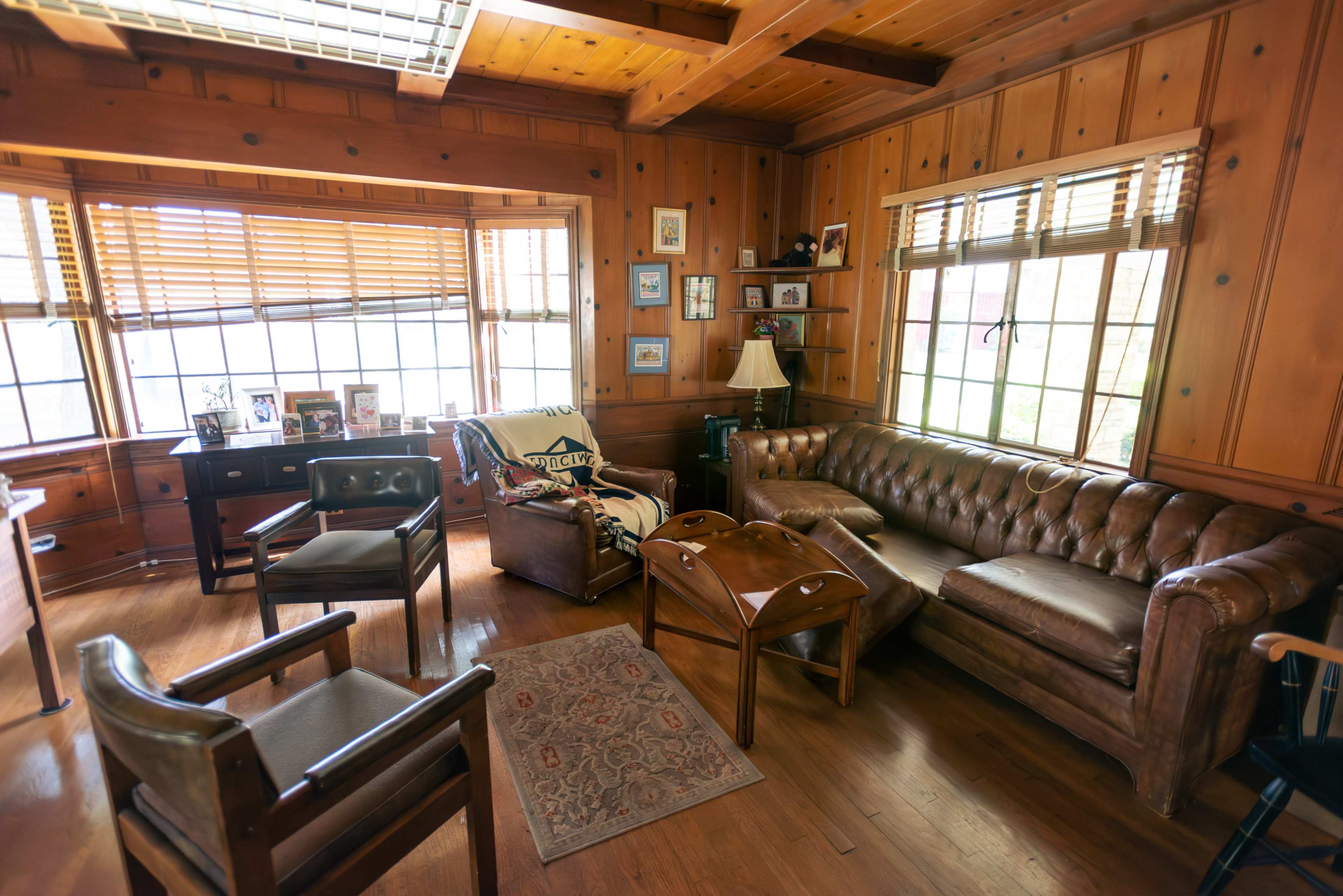 The room features wooden paneled walls, a leather chesterfield sofa, and a mix of wooden furniture, including chairs and side tables.