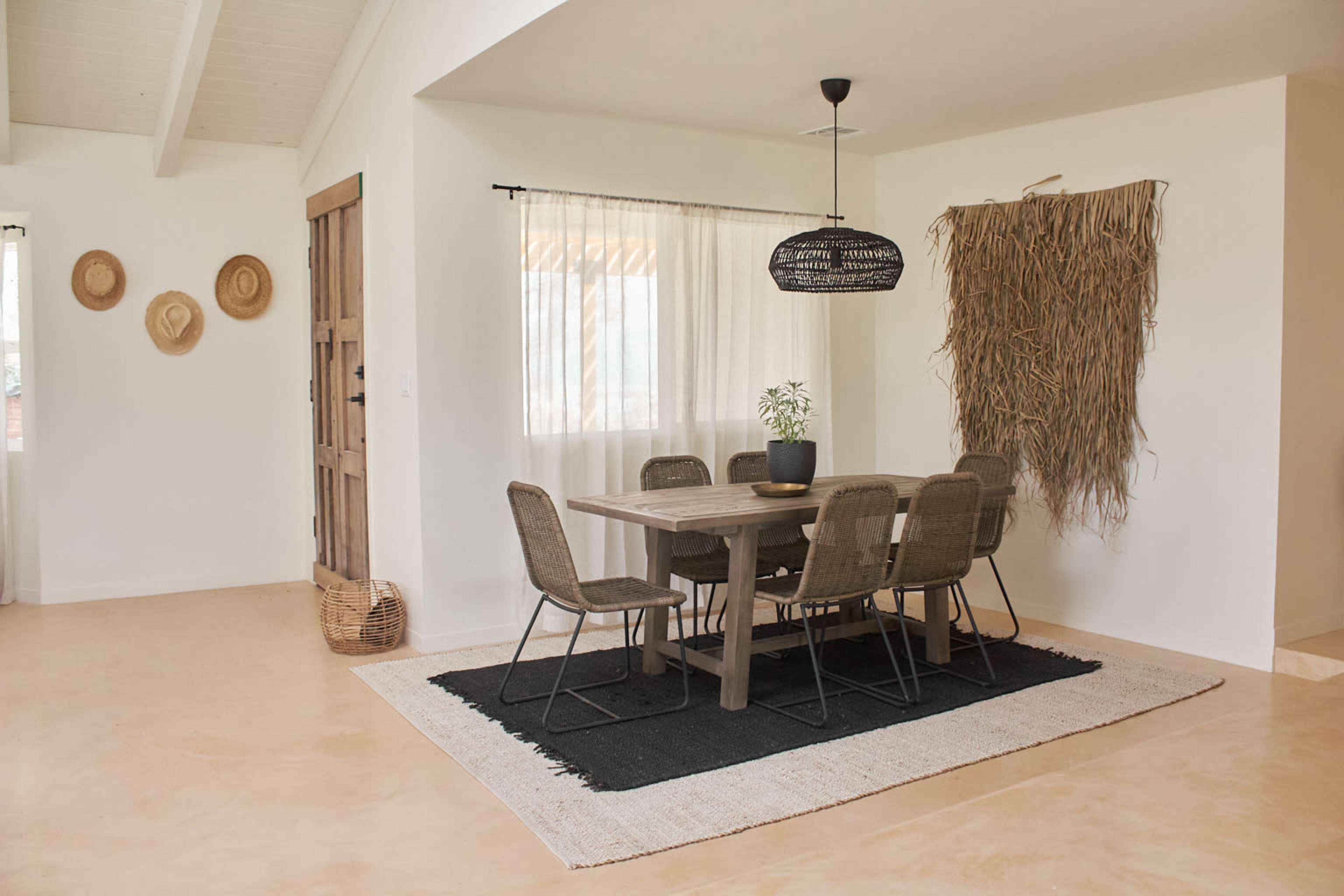 A dining area features a wooden table surrounded by six chairs, with a woven pendant light overhead and decorative elements like wall hangings and hats.