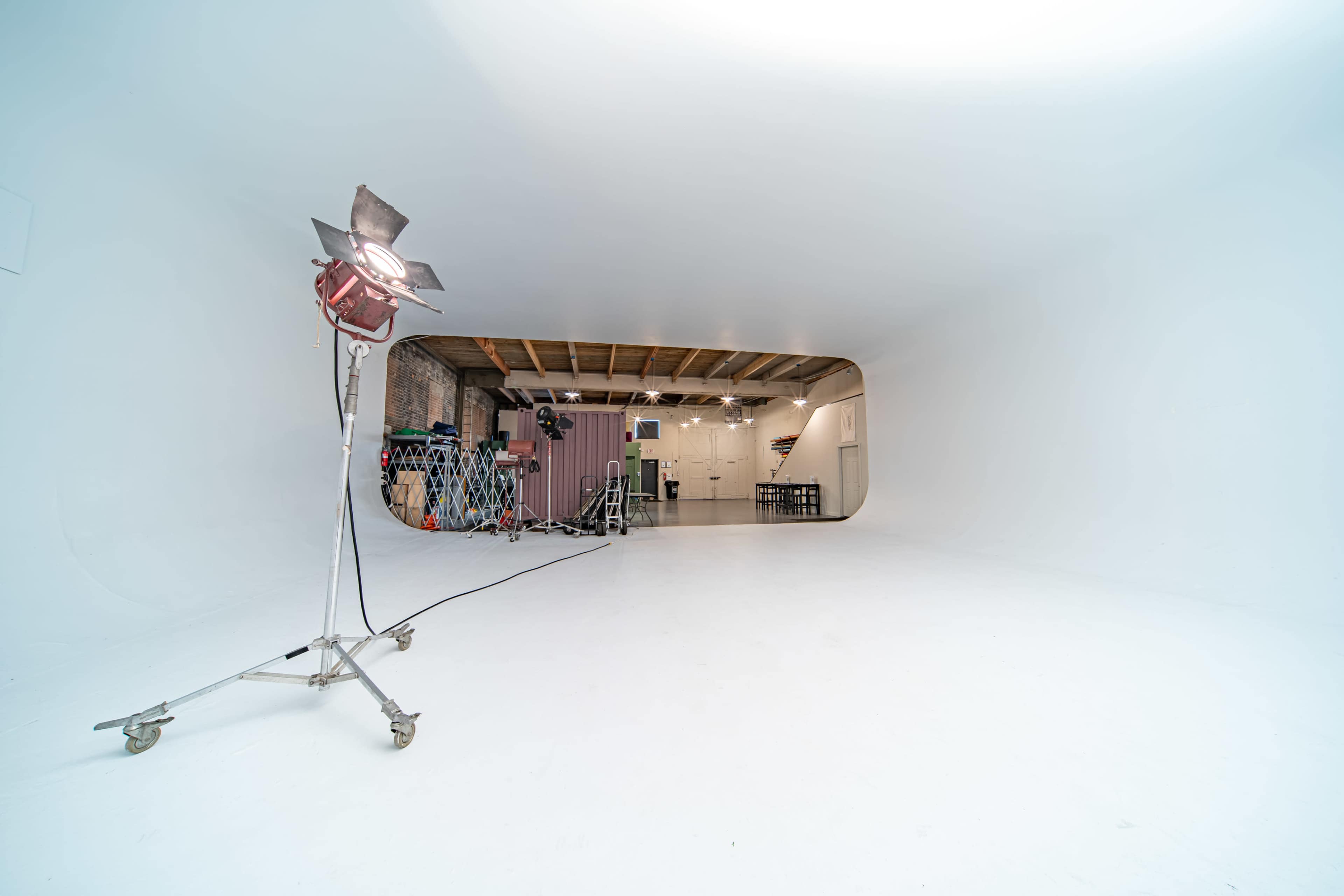 The image shows a spacious, white studio with a lighting fixture on a stand in the foreground and various equipment visible in the background.