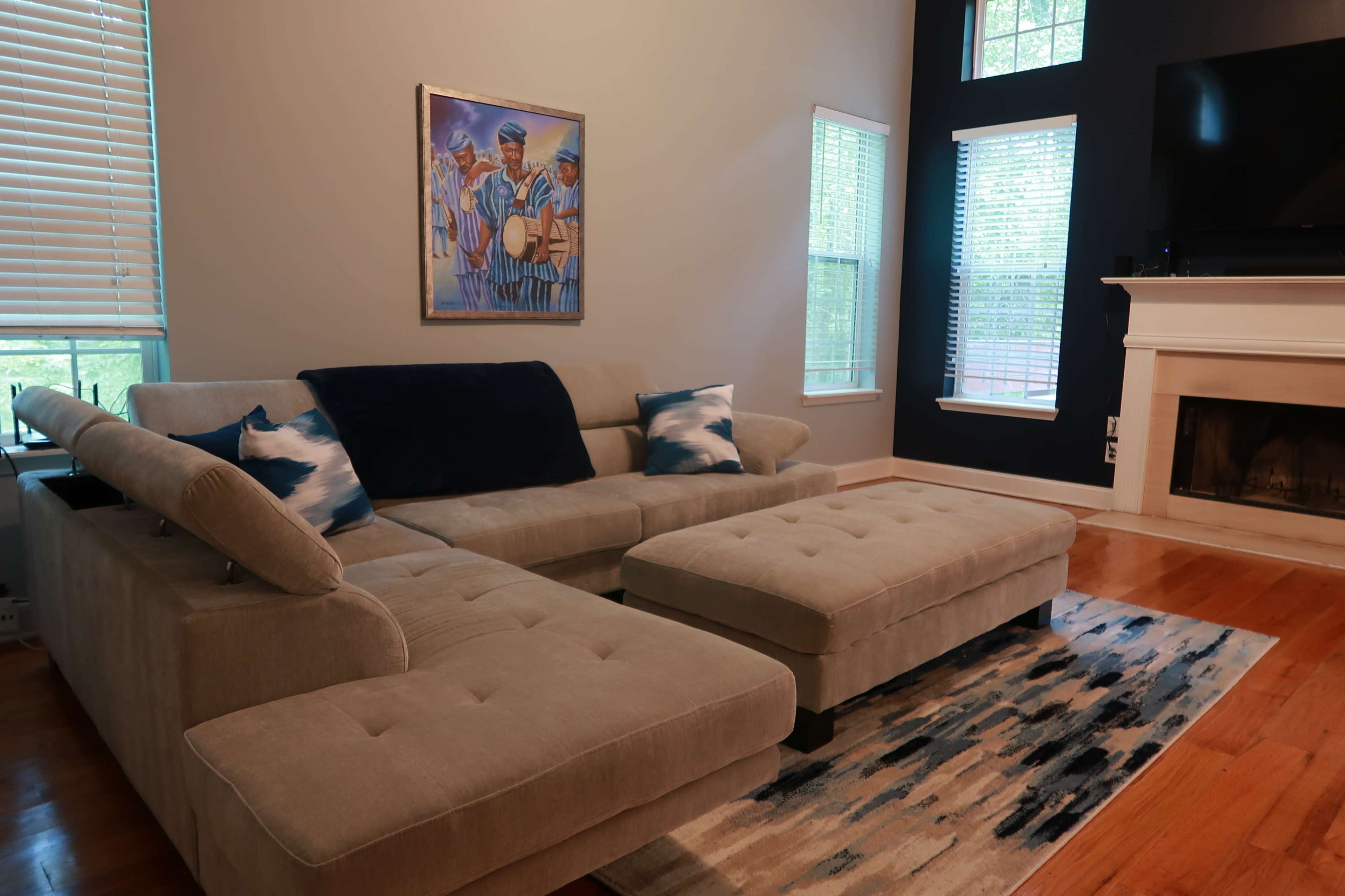 A light-colored sectional sofa with an ottoman sits on a patterned rug in a well-lit living room, featuring large windows and a mounted television.