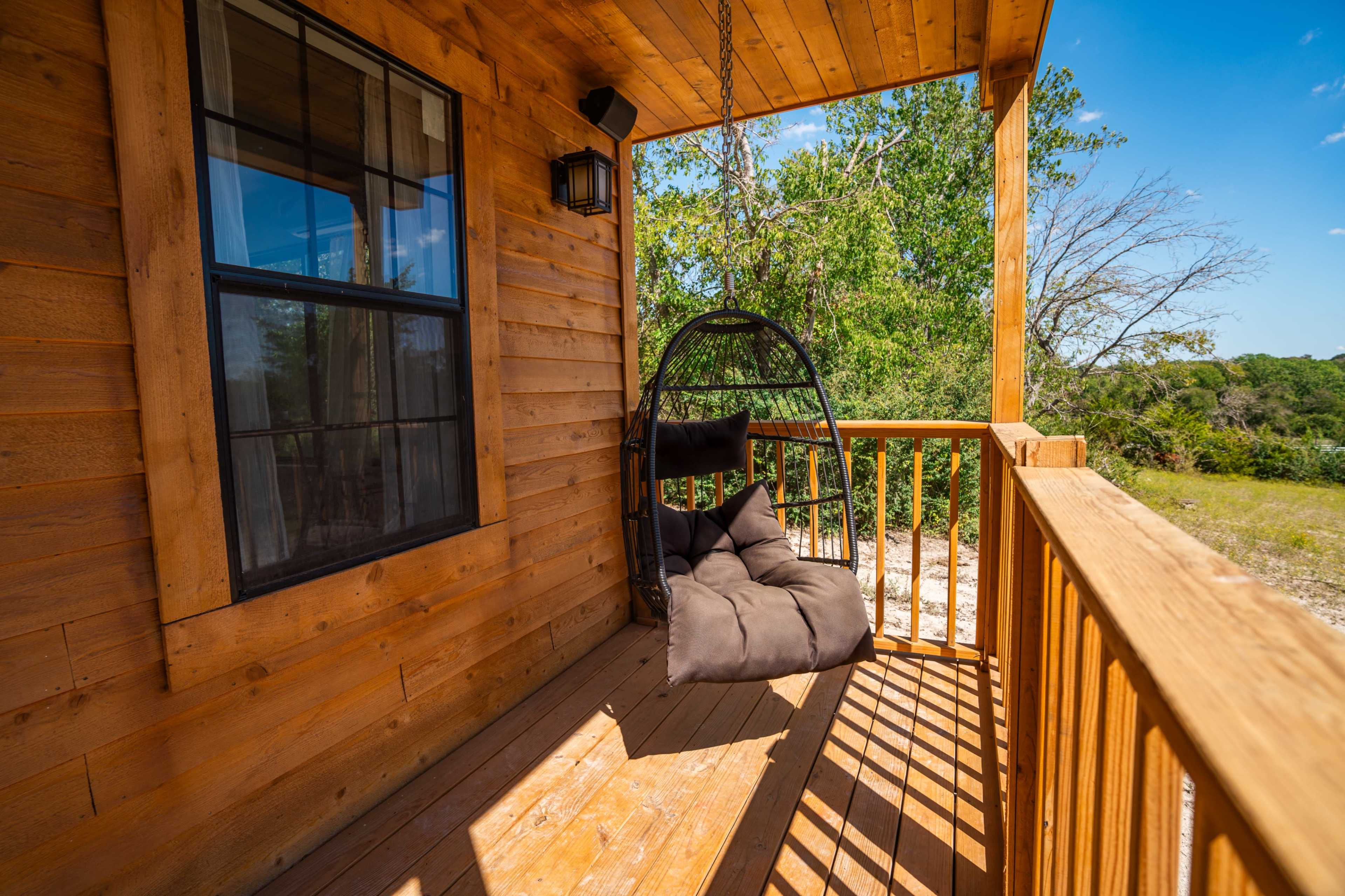 A porch features a hanging chair and wooden railing surrounded by greenery.