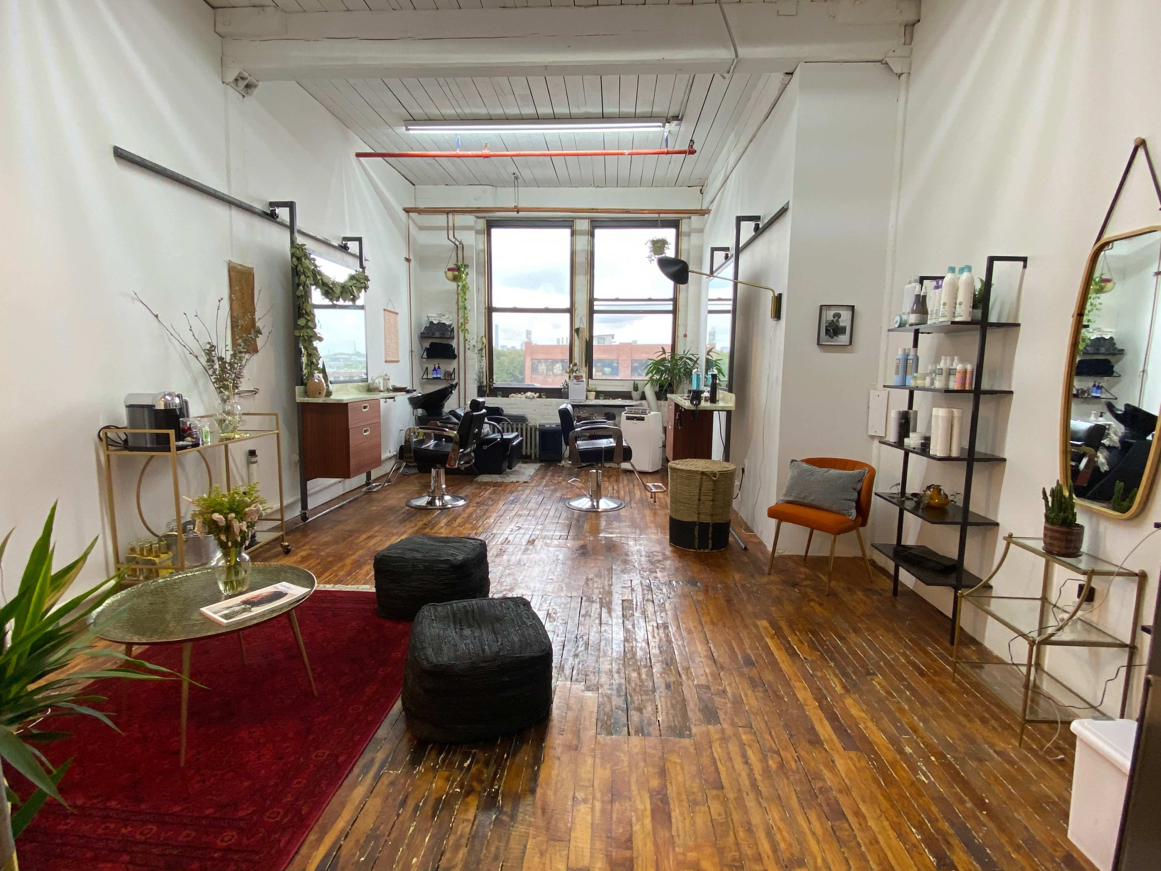 A spacious salon with wooden floors, a seating area, mirrors, shelves displaying products, and styling stations near large windows.