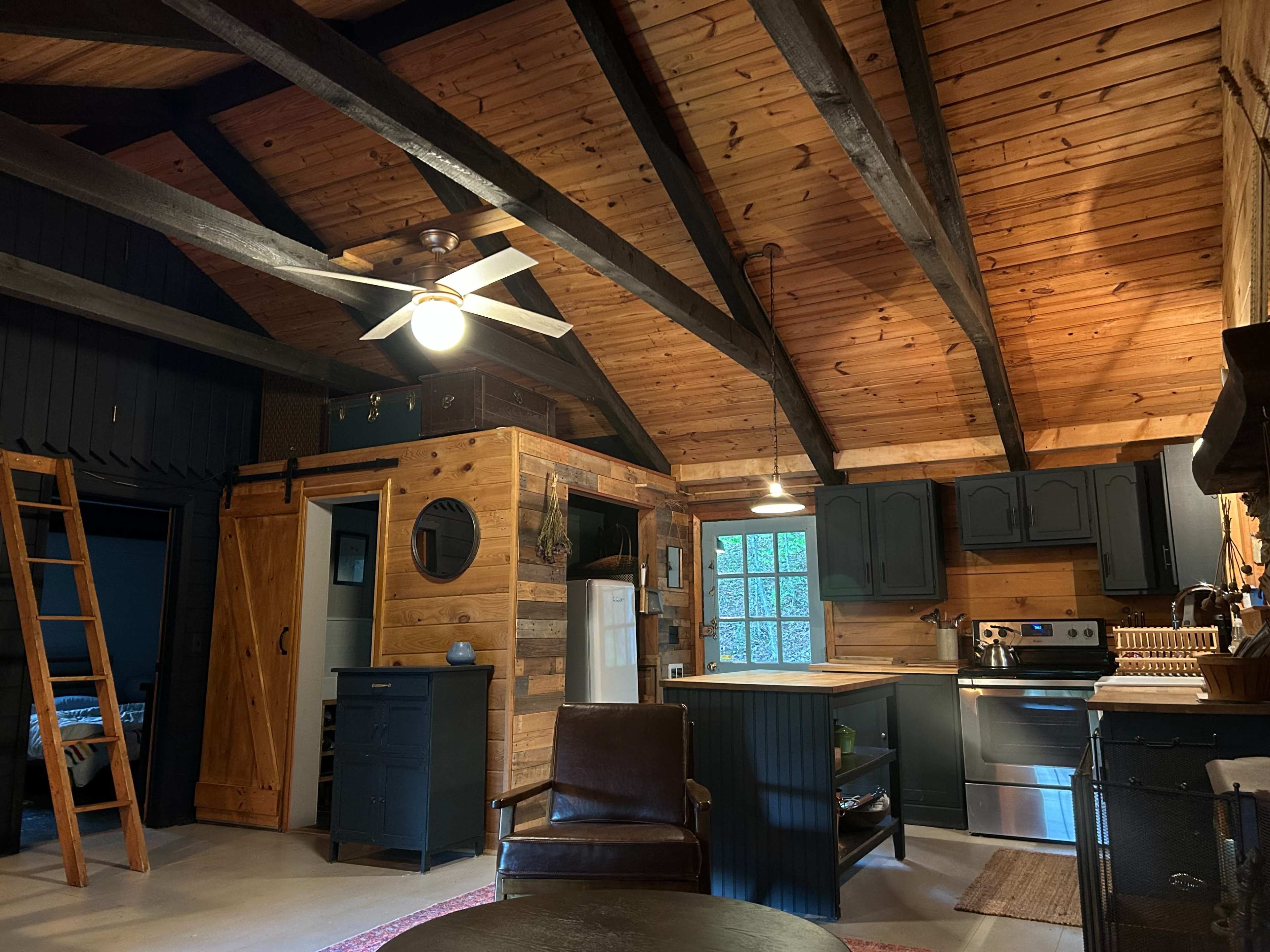 The image shows the interior of a rustic cabin featuring wooden beams, a kitchen area with dark cabinets and a stainless steel stove, and a cozy seating area with a leather chair.