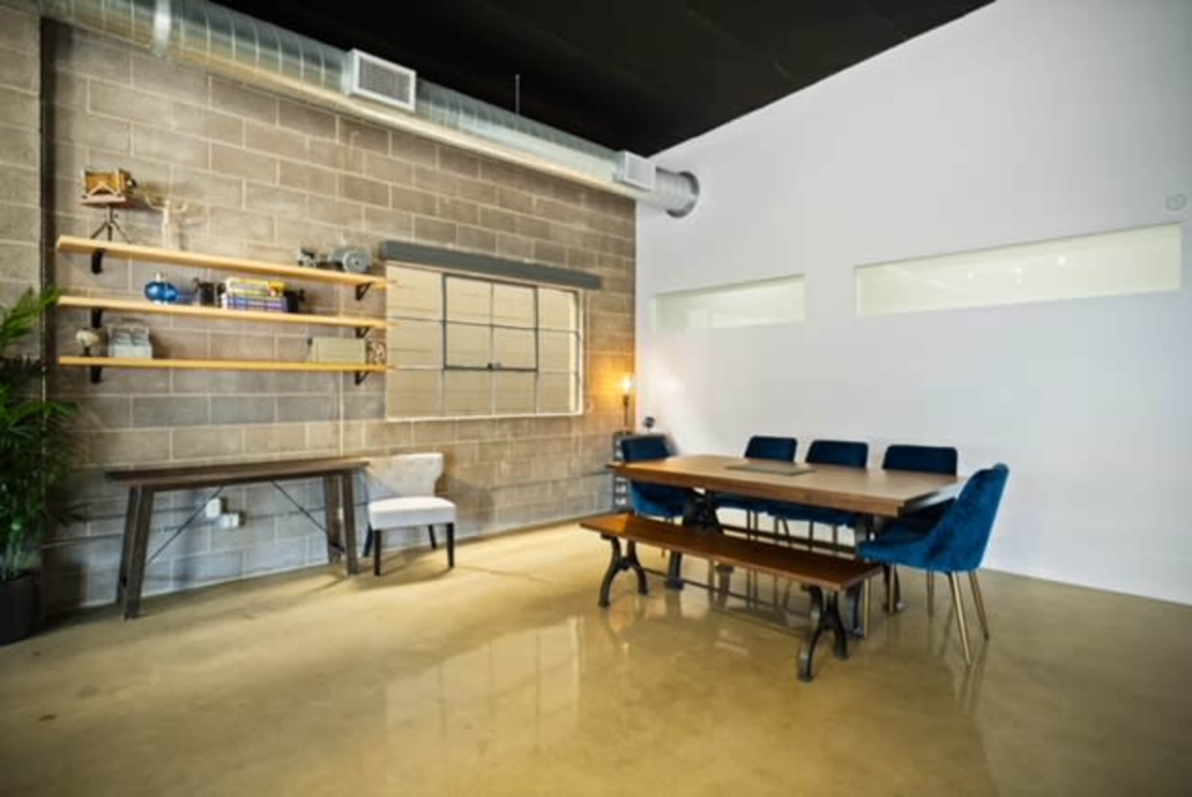 A modern meeting room features a wooden table surrounded by blue chairs, with a concrete wall adorned by shelves and a window providing natural light.