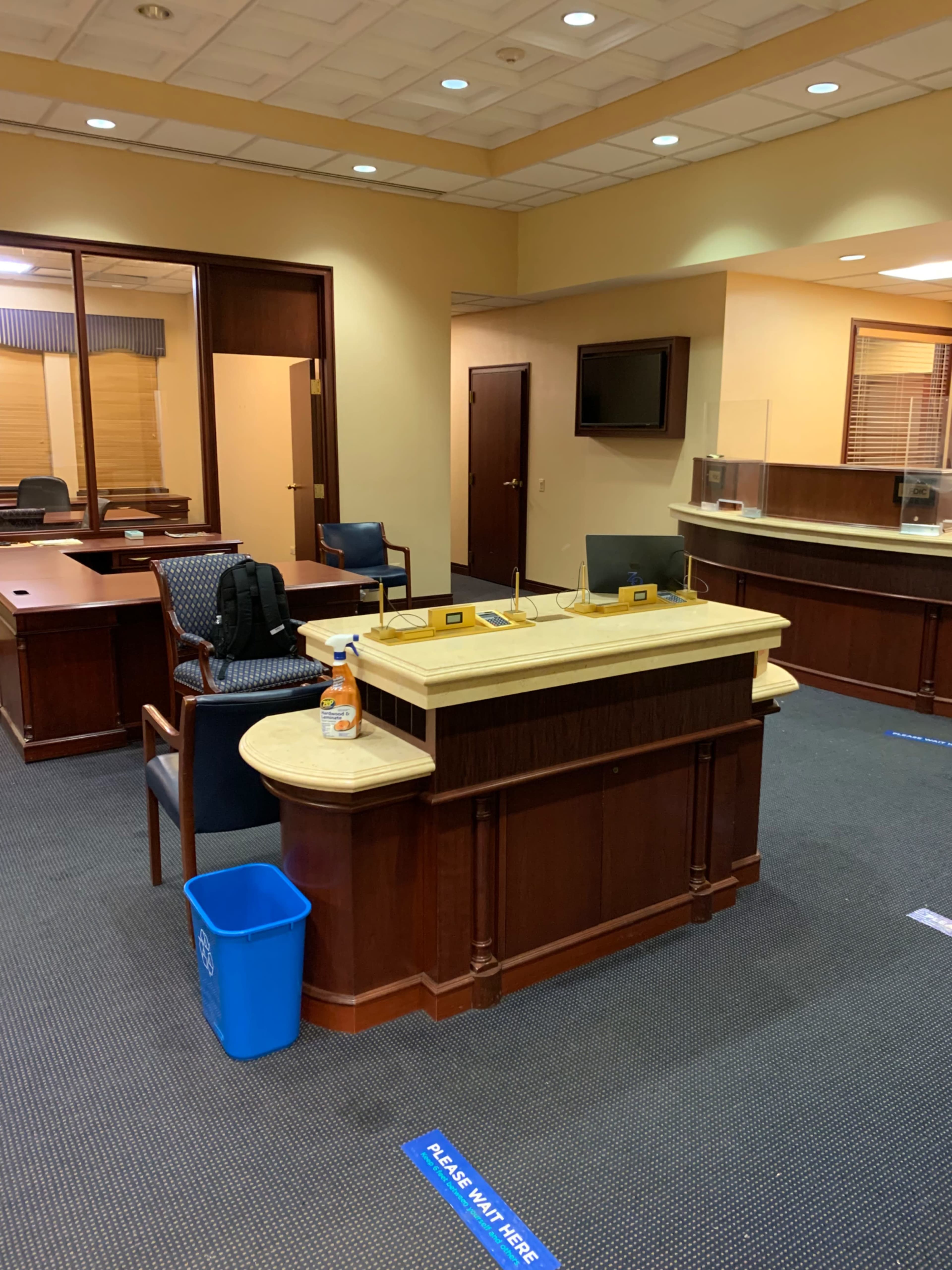 The image shows a partially furnished office space with a reception desk, a chair, and a wastebasket, featuring signs on the floor indicating where to stand.