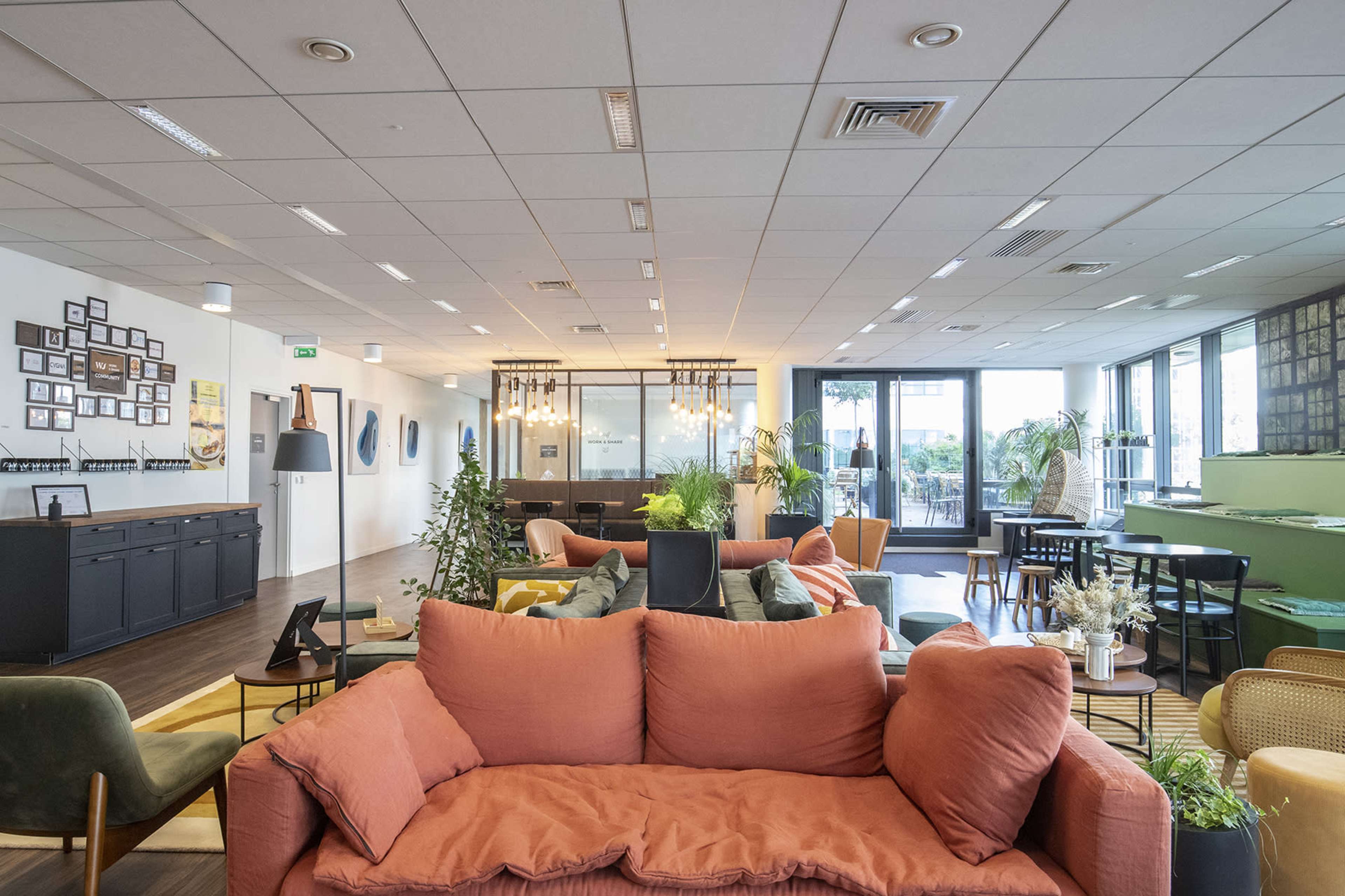 A modern, open office space with various seating areas, plants, and large windows allowing natural light.