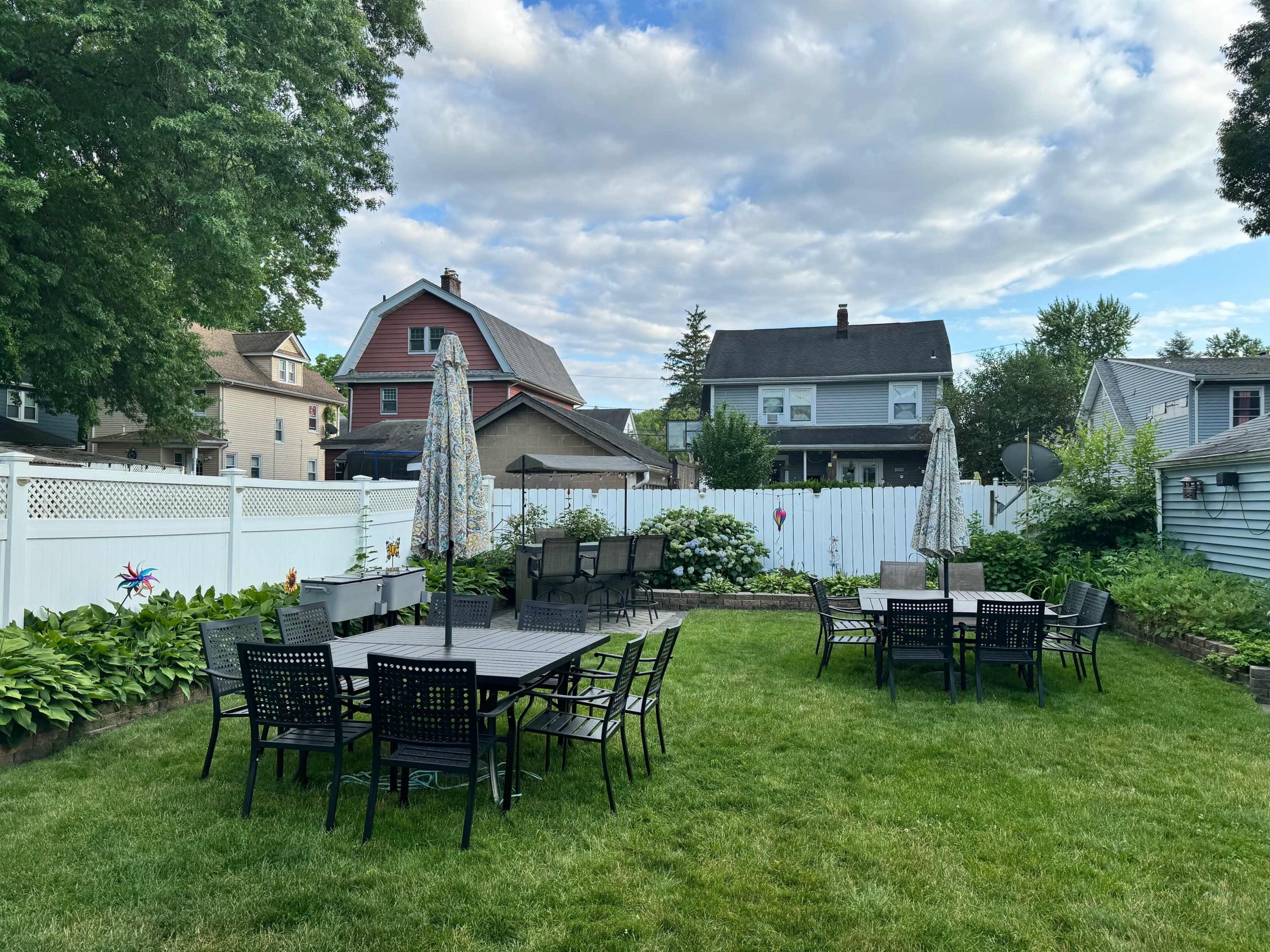 A backyard with several black patio tables and chairs arranged on a green lawn, surrounded by a white fence and various houses in the background.