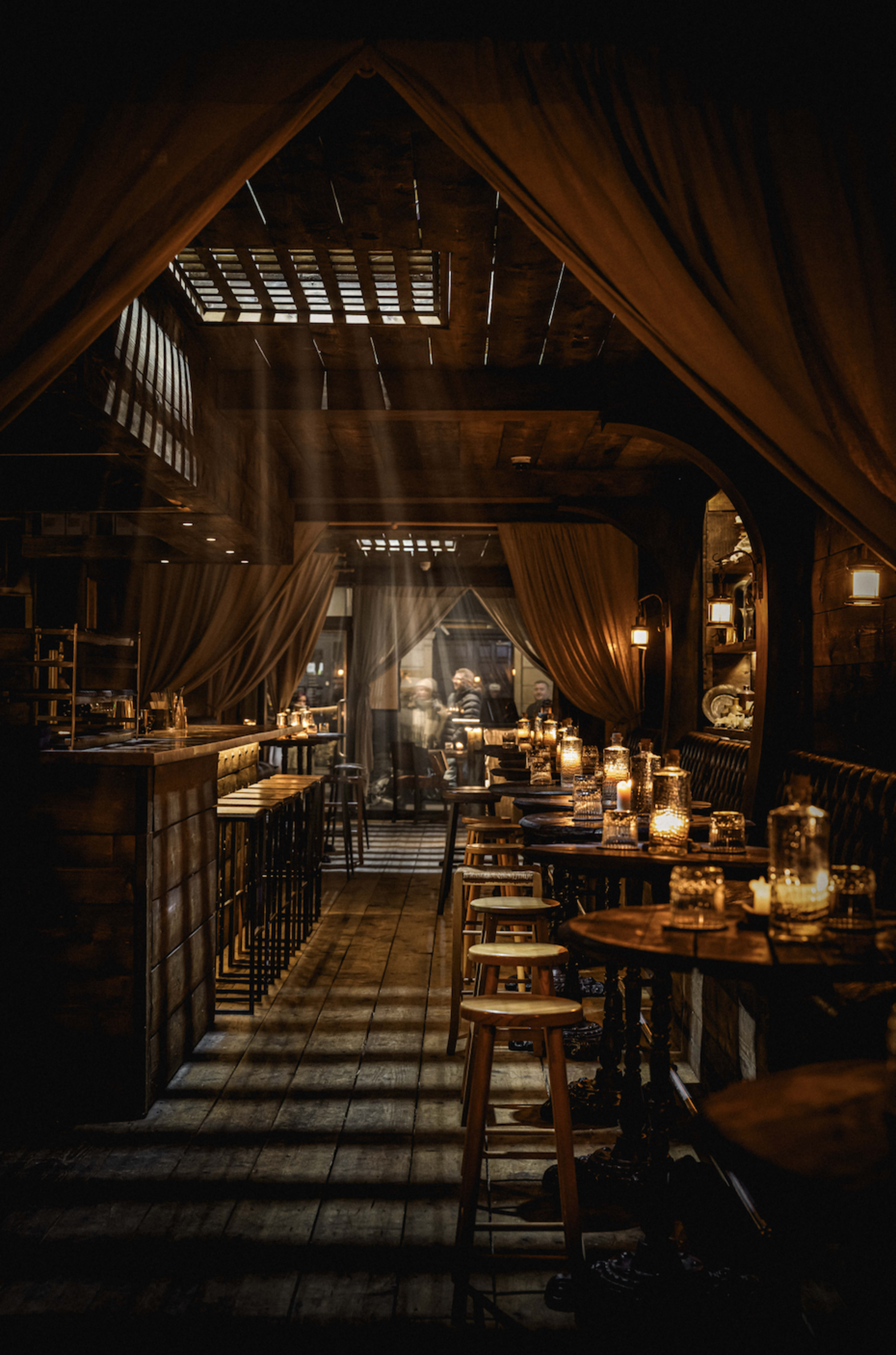 The interior of a dimly lit restaurant features wooden beams, soft drapery, and tables illuminated by flickering candlelight.